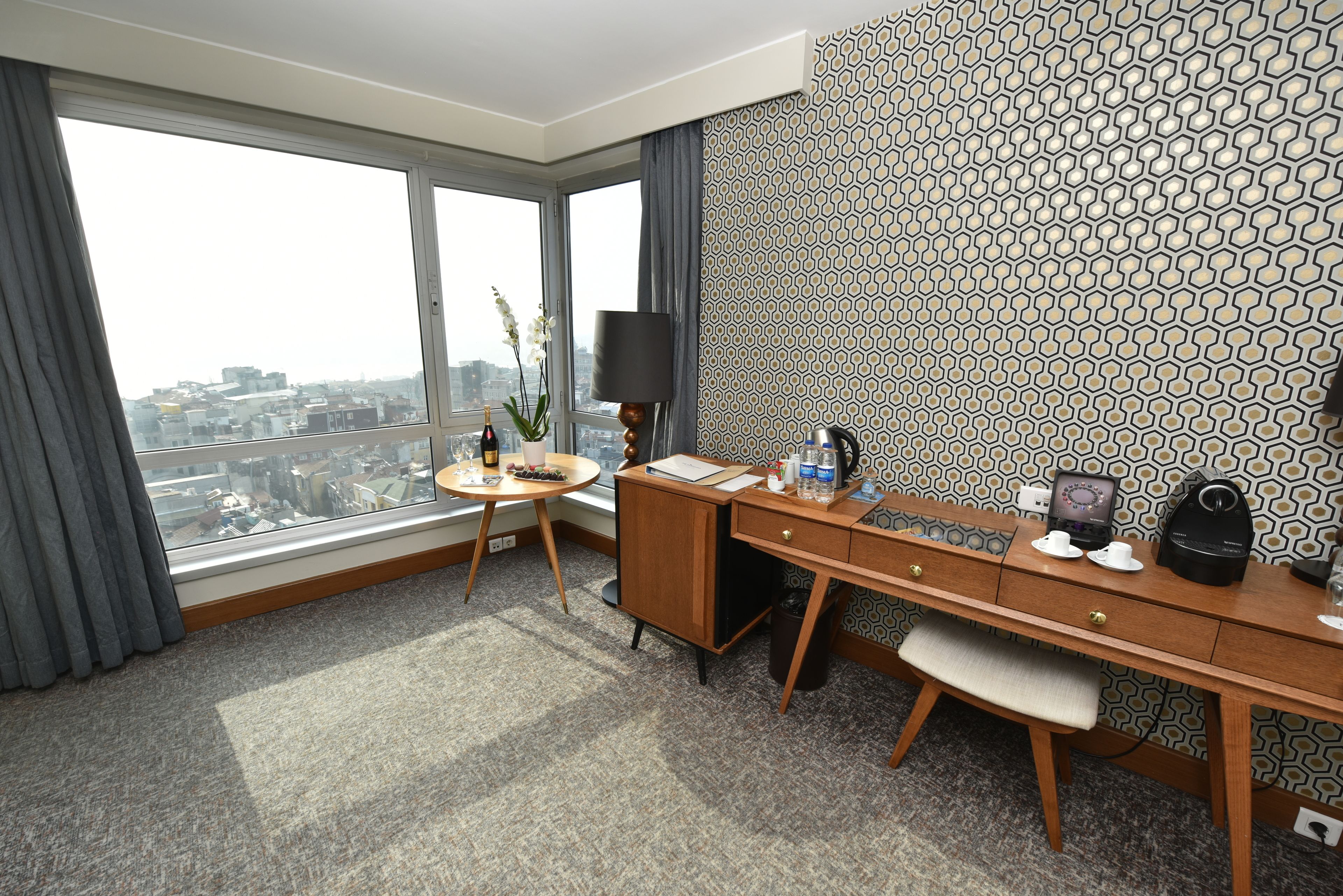 A hotel room with a large window, a desk, a chair, a small table, a lamp, a vase with flowers, and a patterned wallpaper.