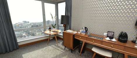 Habitación de hotel con un ventanal grande, un escritorio, una silla, una mesita, una lámpara, un jarrón con flores y un papel pintado con estampado.