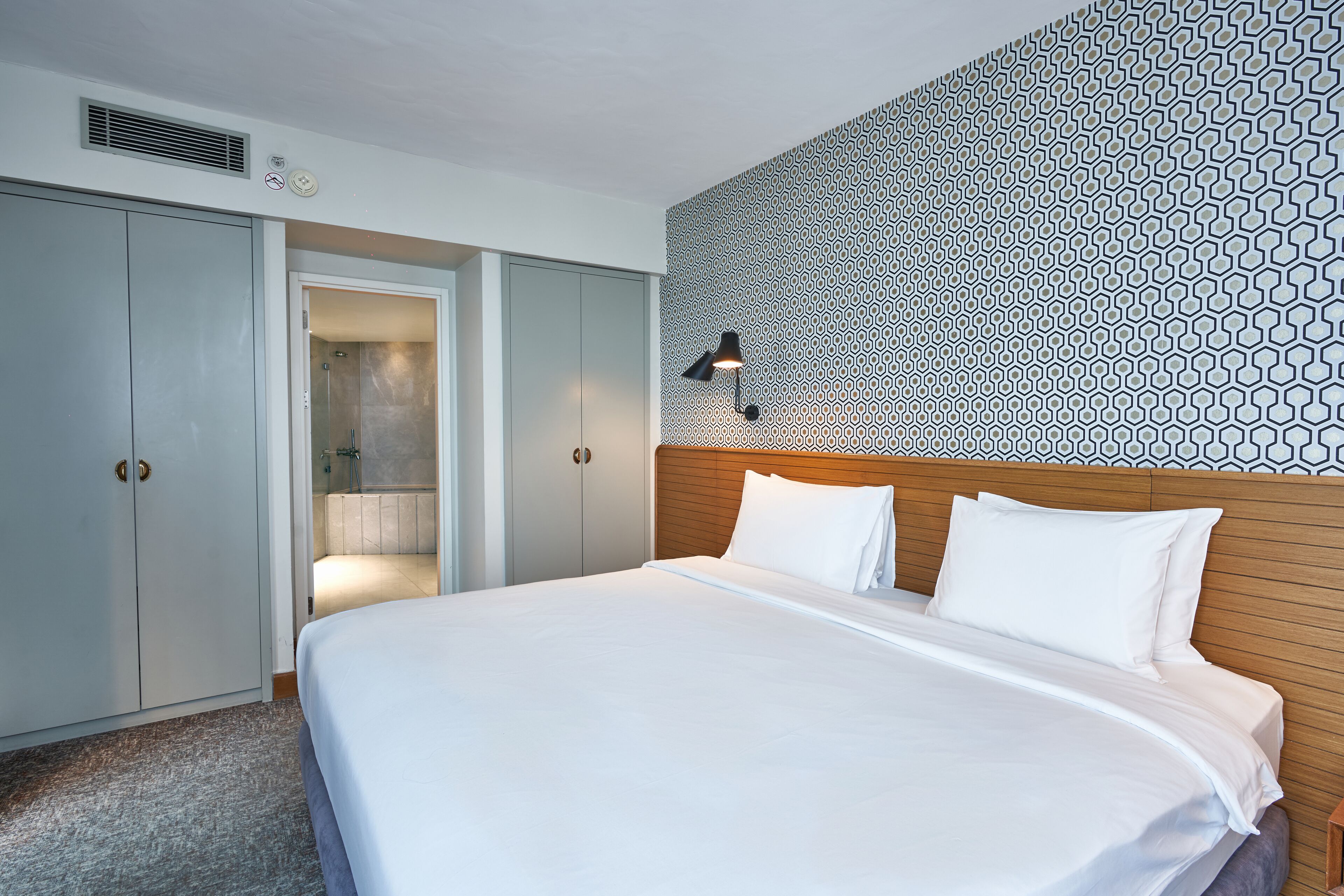A hotel room with a large bed, a wooden headboard, a patterned wall, a wardrobe, and a bathroom with a shower.
