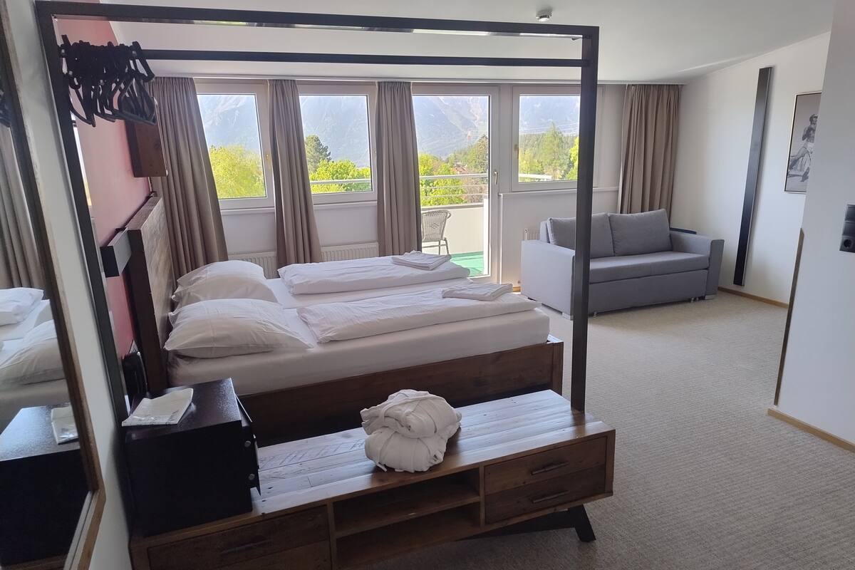 A hotel room with a four-poster bed, a grey sofa, a wooden nightstand, and a view of mountains through large windows.