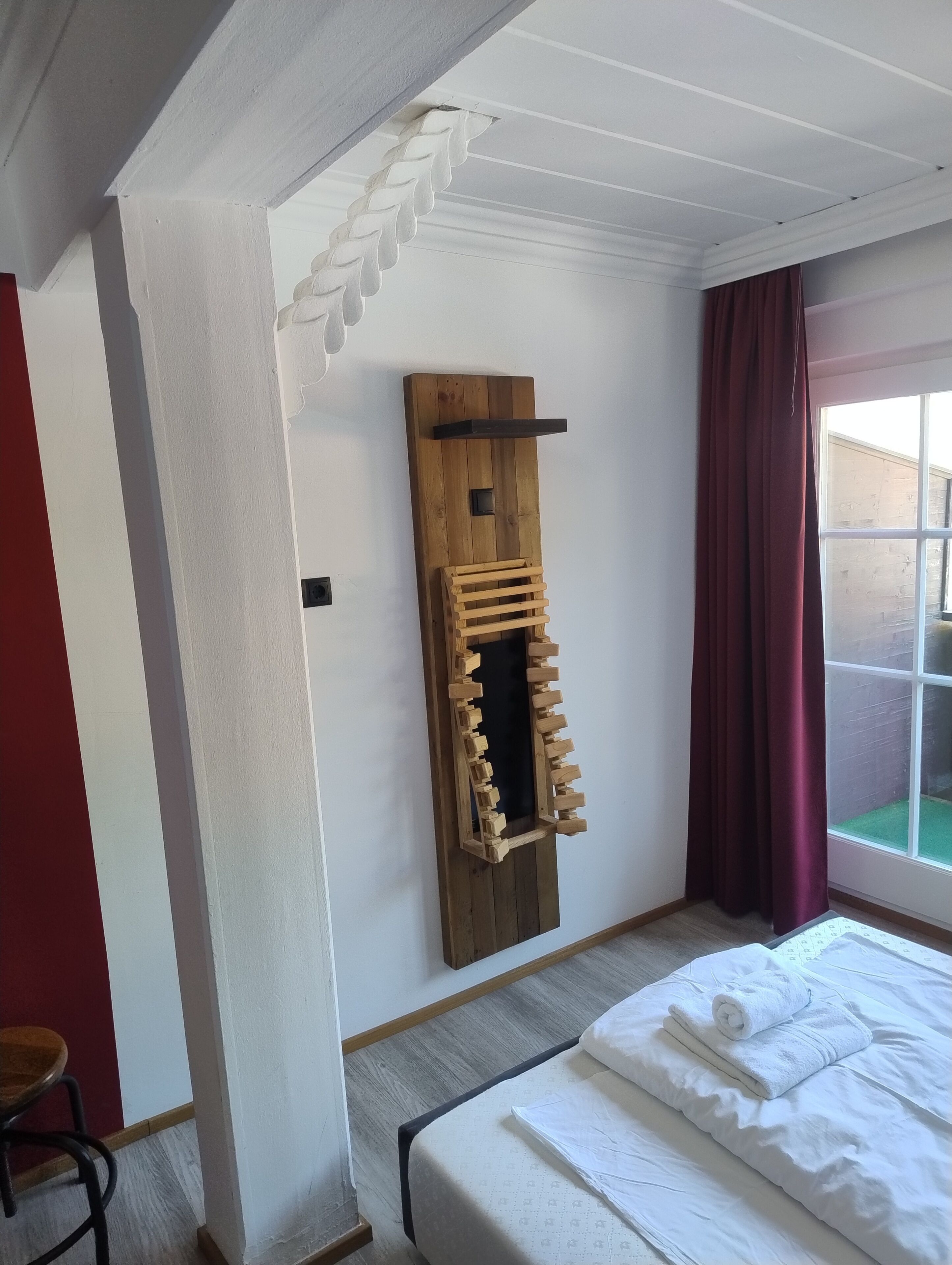 Ein Schlafzimmer mit einem Bett, einem Holzregal, einem Fenster mit Vorhängen und Blick auf eine Ziegelwand.