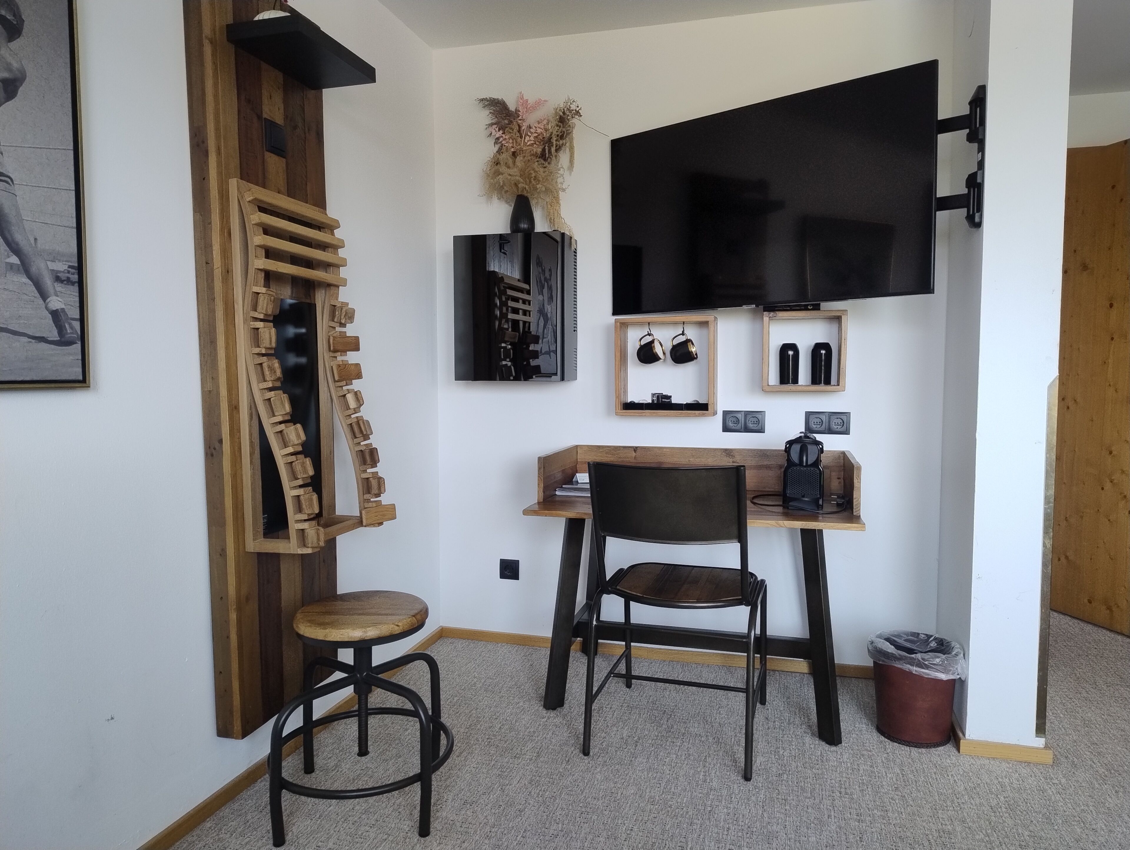 Una habitación pequeña y acogedora con un escritorio de madera, dos sillas, un televisor montado en la pared y un jarrón decorativo con flores secas.