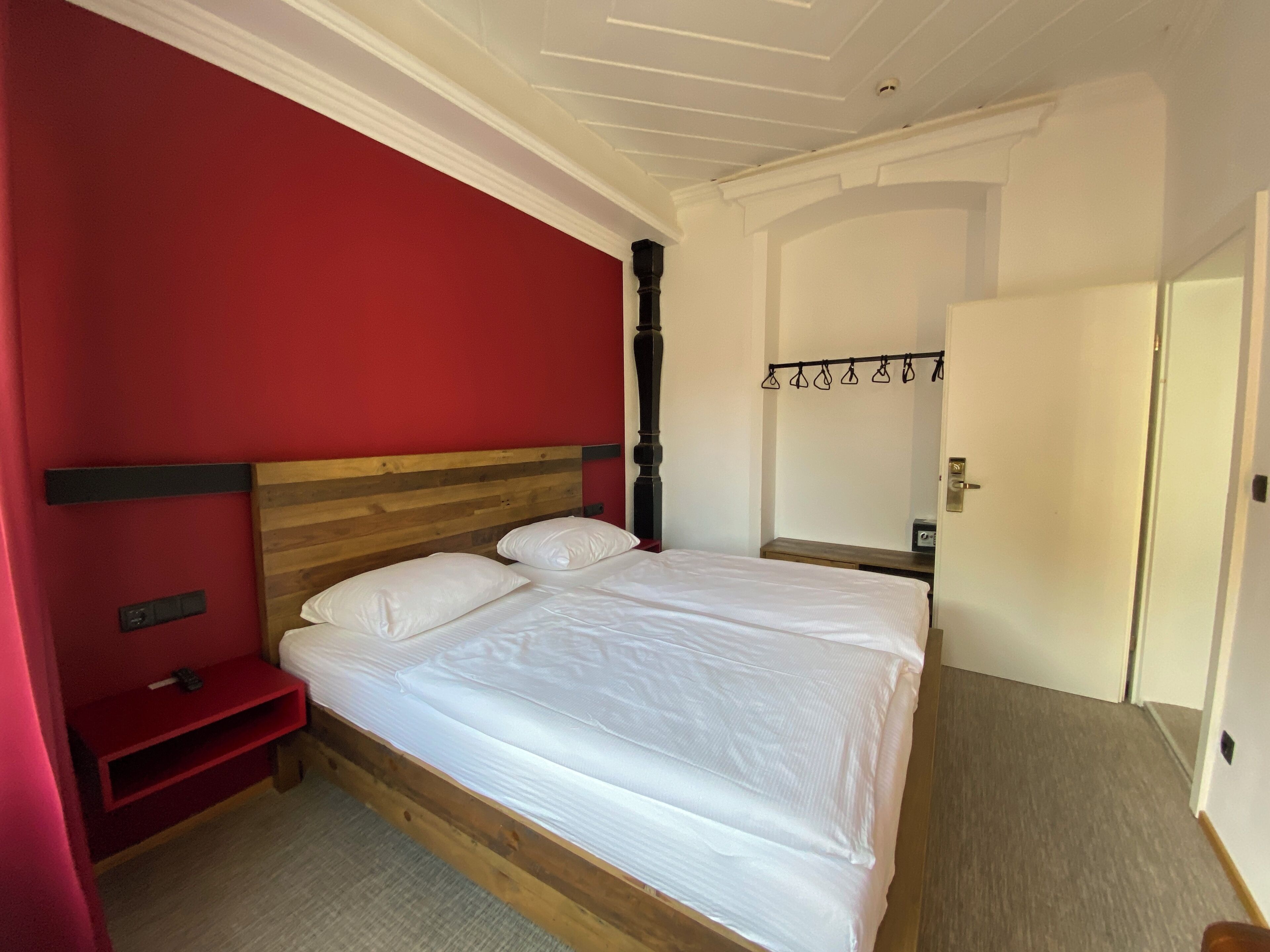 A room with a bed, a red wall, a wooden headboard, a small bedside table, and a door.