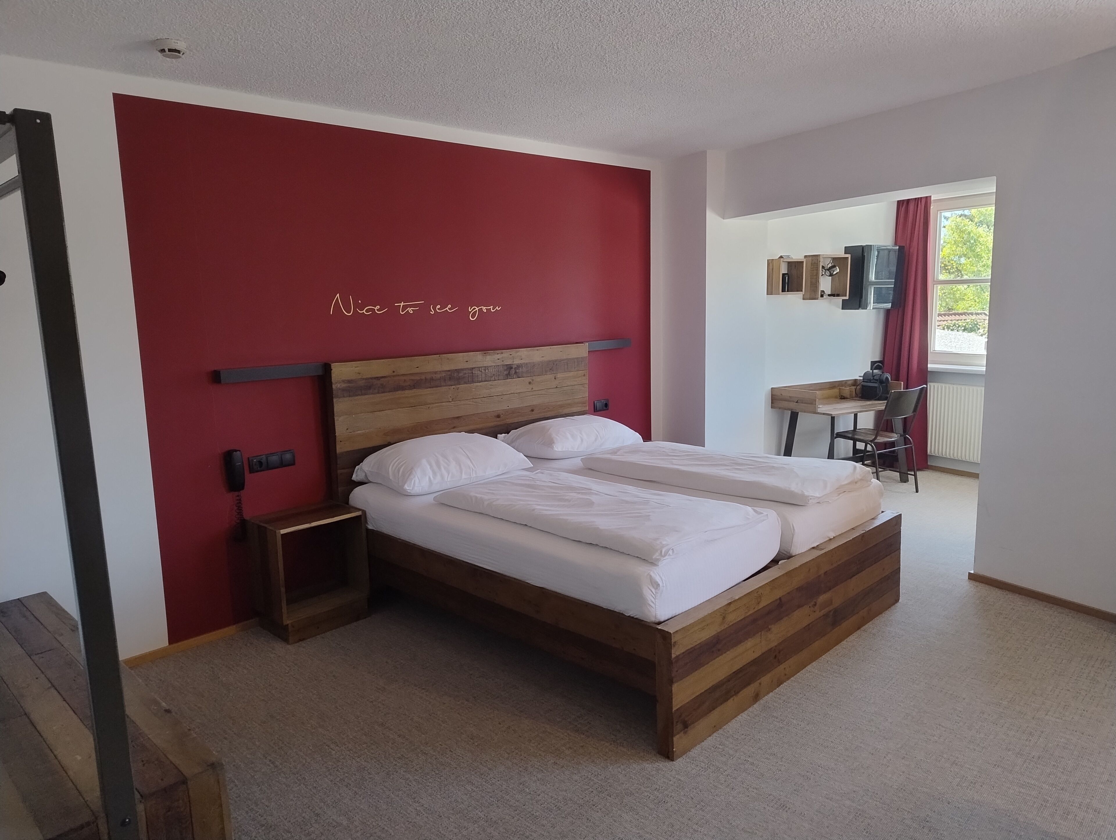 Une chambre avec un lit en bois, un mur d’accent rouge, une petite table de chevet et une fenêtre donnant sur un beau paysage.