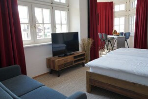 A hotel room with a bed, a TV on a wooden stand, a desk with chairs, and large windows with red curtains.