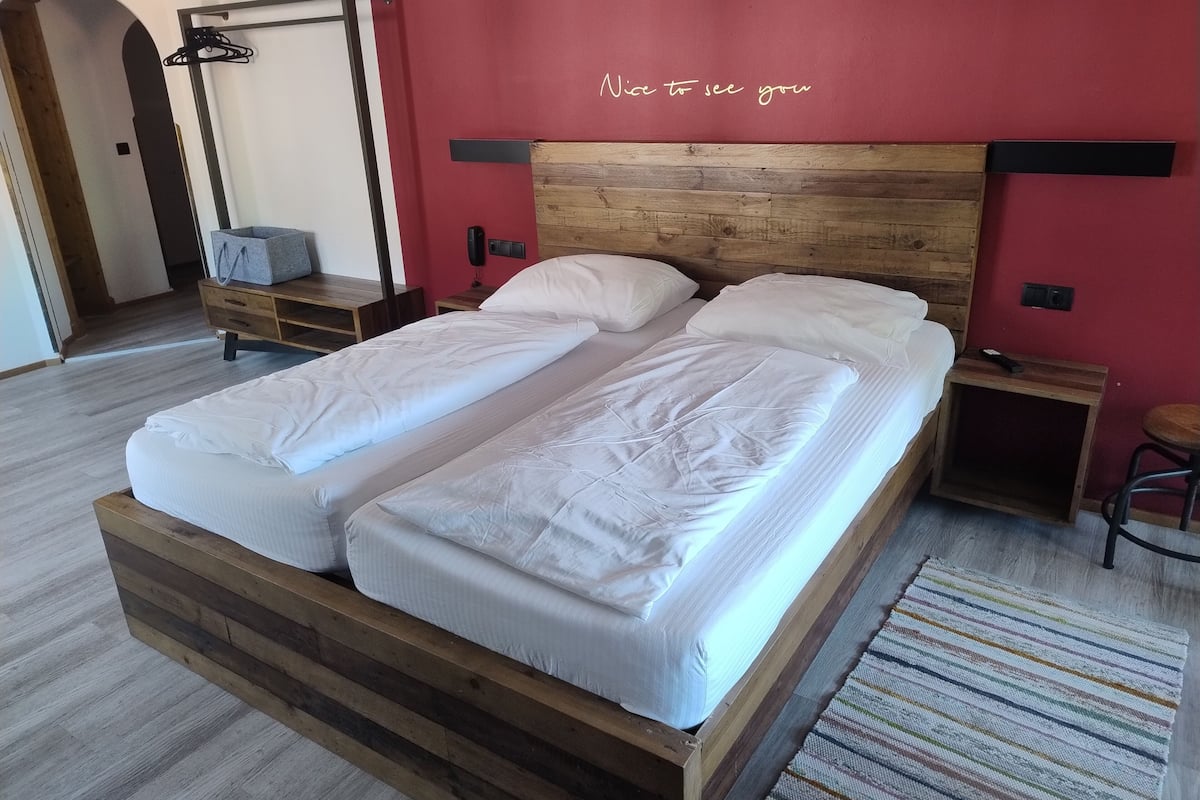 A double bed with a wooden headboard, two bedside tables, a hanging coat rack, and a red wall with a 'Nice to see you' sign.