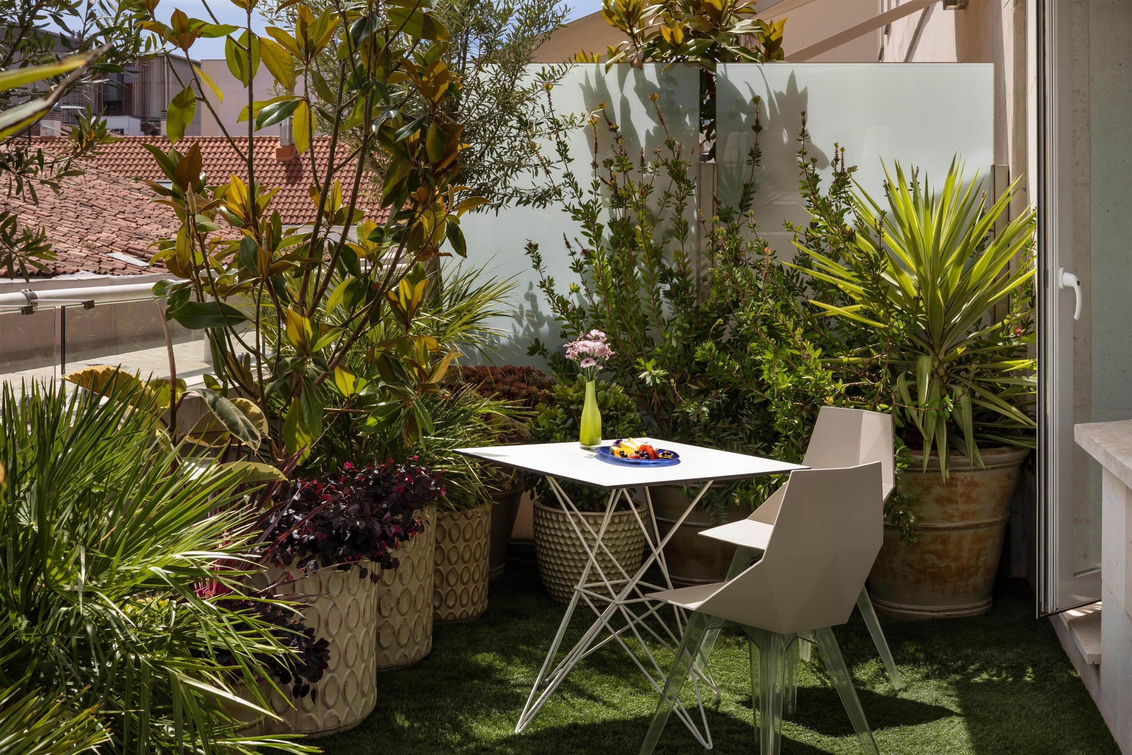 Une petite terrasse avec une table et des chaises, entourée de plantes en pot et offrant une vue sur un immeuble.