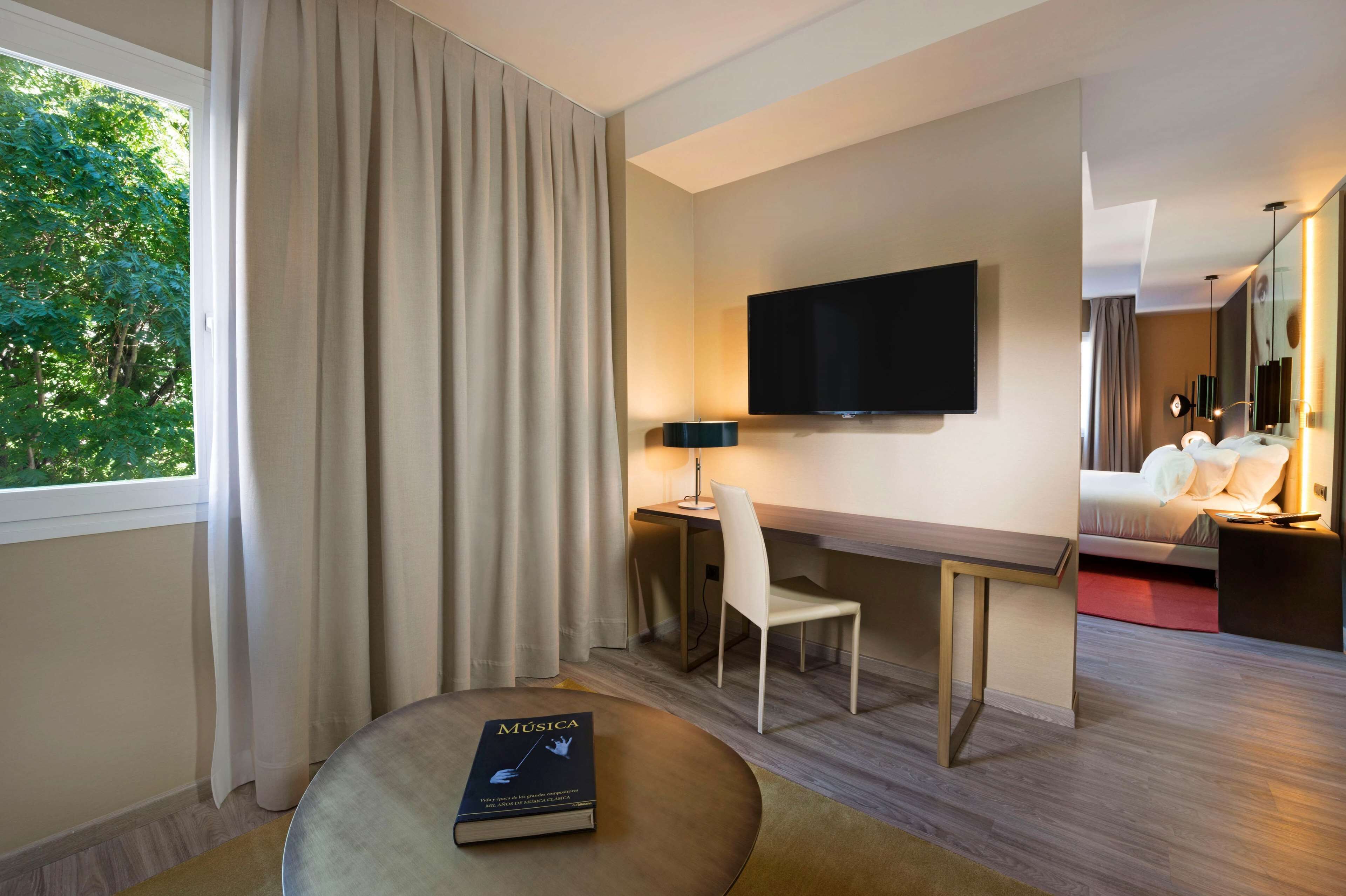 A hotel room with a desk, chair, TV, bed, and a book titled "MUSICA" on a round table.