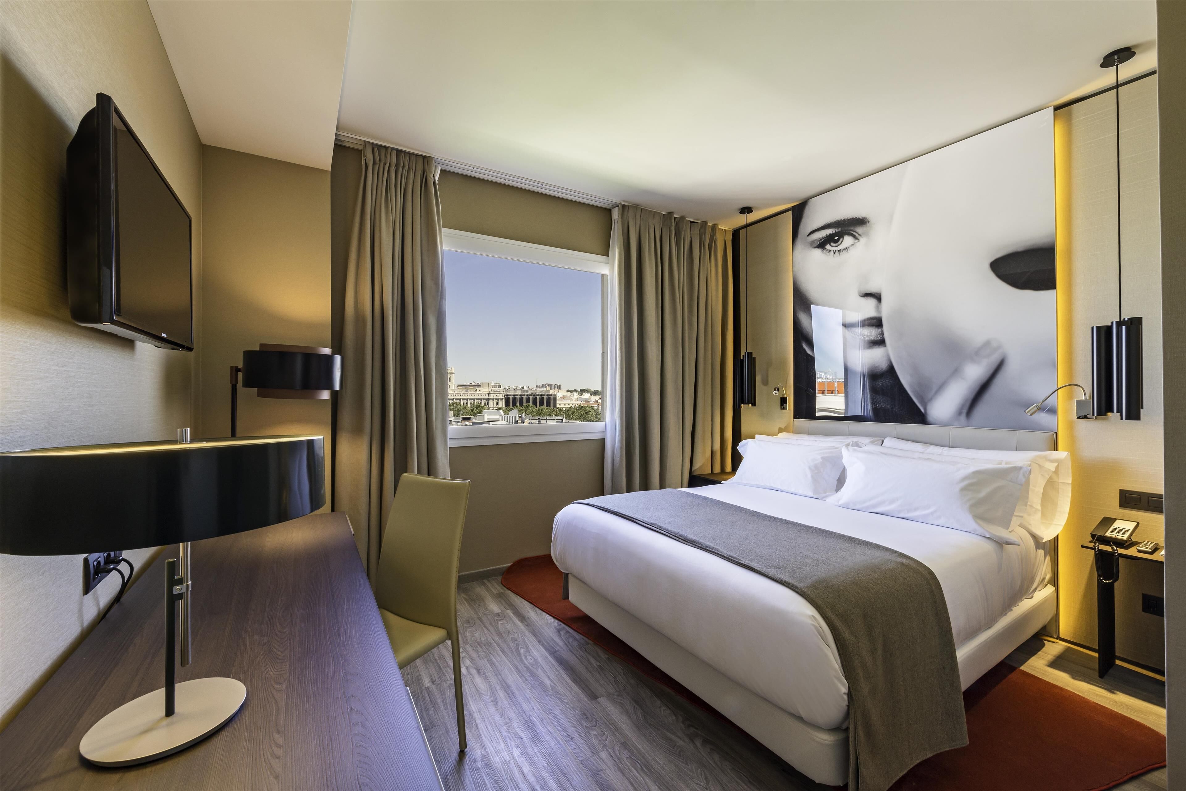 A modern hotel room with a large bed, a desk, a flat-screen TV mounted on the wall, and a window with curtains.