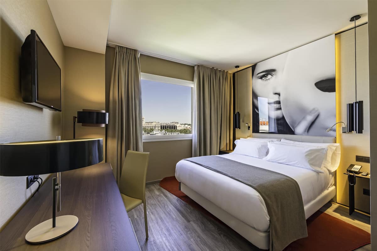 A modern hotel room with a large bed, a desk, a flat-screen TV mounted on the wall, and a window with curtains.
