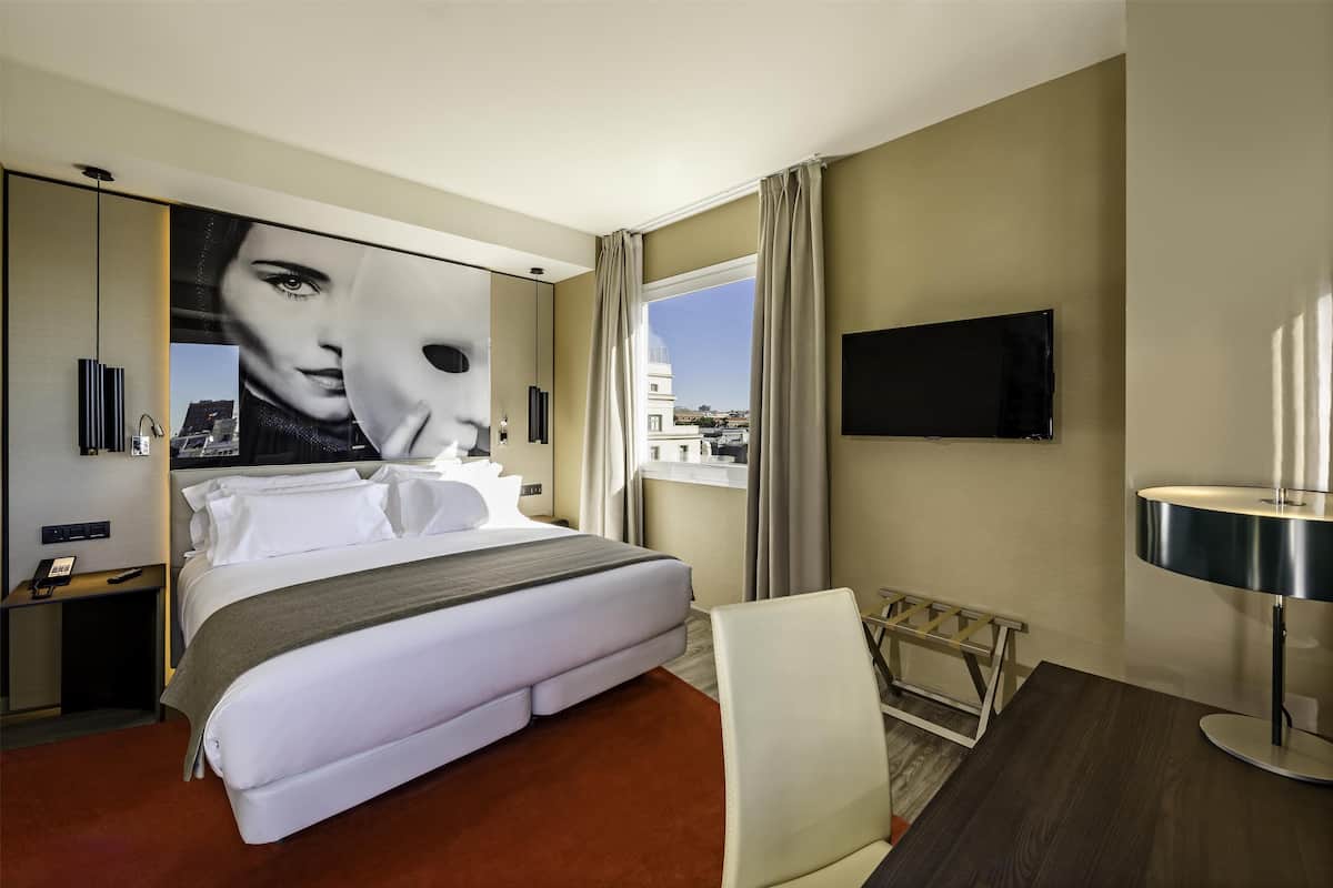 A modern hotel room with a large bed, a desk, a chair, a TV, and a view of the cityscape.