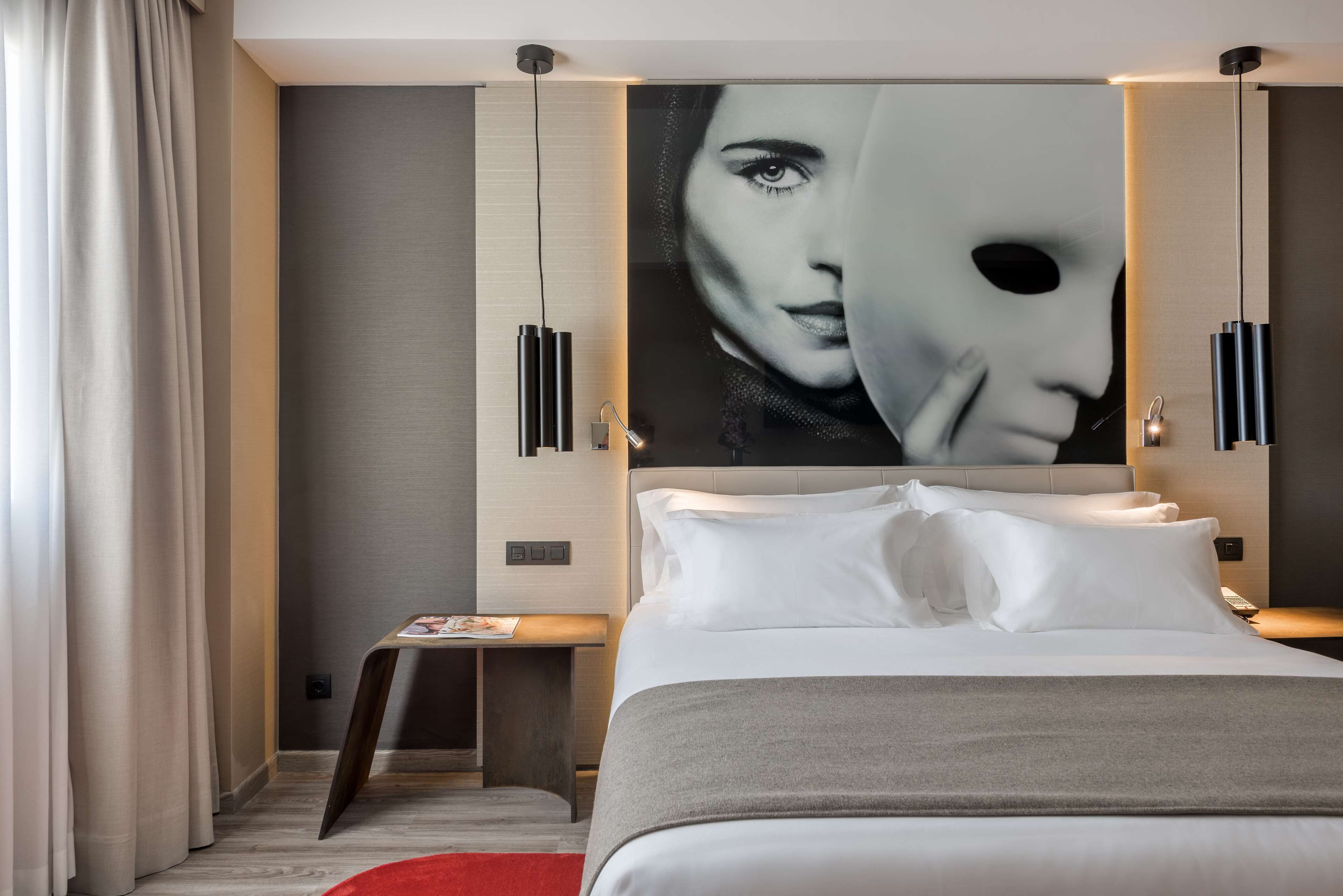 Una habitación de hotel moderna con una cama grande, una mesita de noche, una lámpara fijada a la pared y un cuadro abstracto de gran tamaño.