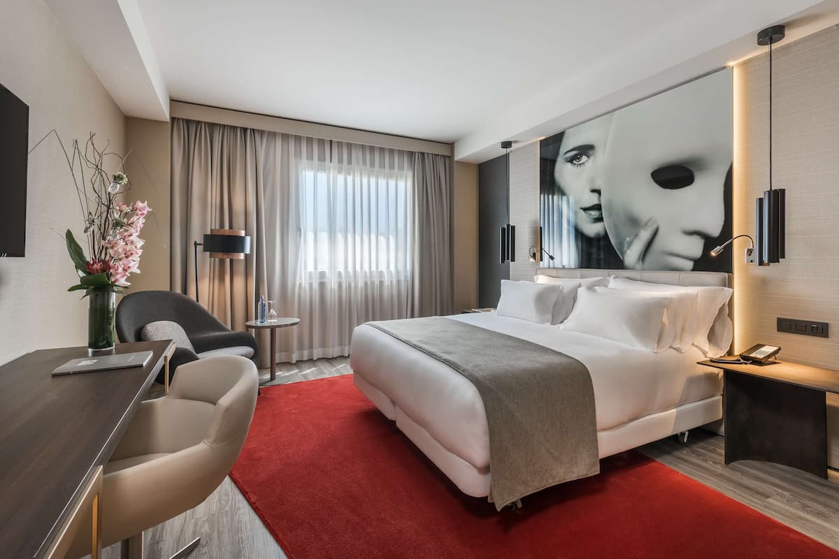 A modern hotel room with a large bed, a desk with a chair, a red carpet, and a large window with curtains.