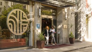 A hotel entrance with a large decorative sign, two potted plants, and a red carpet leading to the entrance. - NH Collection Madrid Suecia (Madrid)
