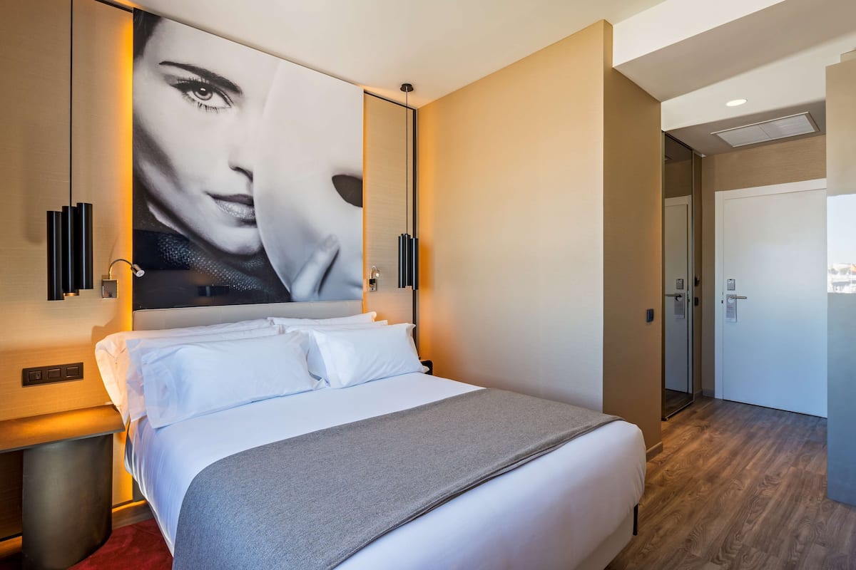 A modern hotel room with a large bed, a bedside table, a wall-mounted artwork, and a door leading to a bathroom.