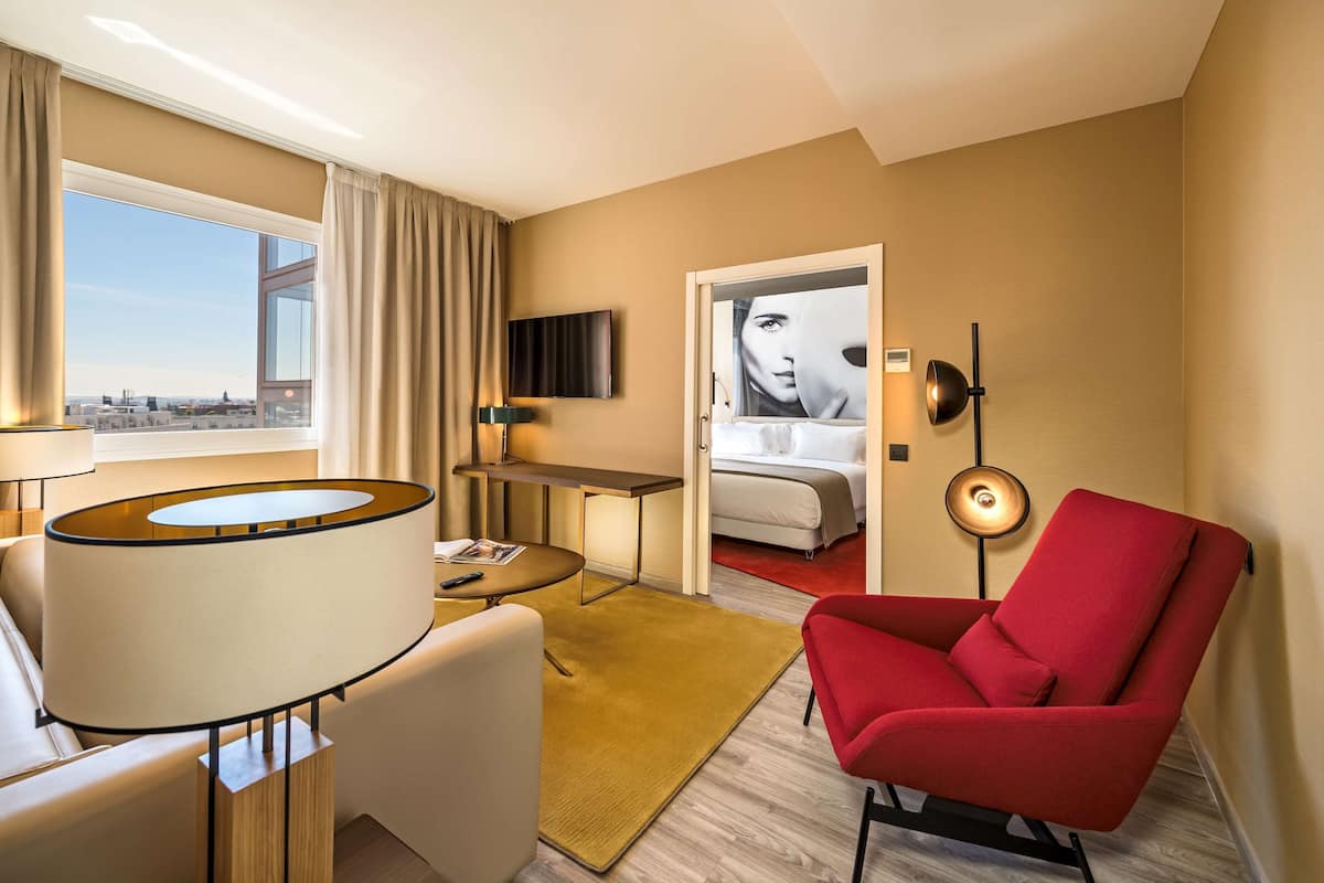 A hotel room with a large window, a red armchair, a sofa, a small table, a TV, and a bed with a view of the city.
