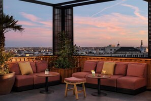 A rooftop terrace with a city view, featuring a wooden deck, red cushioned seating, and a small table with drinks. - NH Collection Madrid Suecia (Madrid)