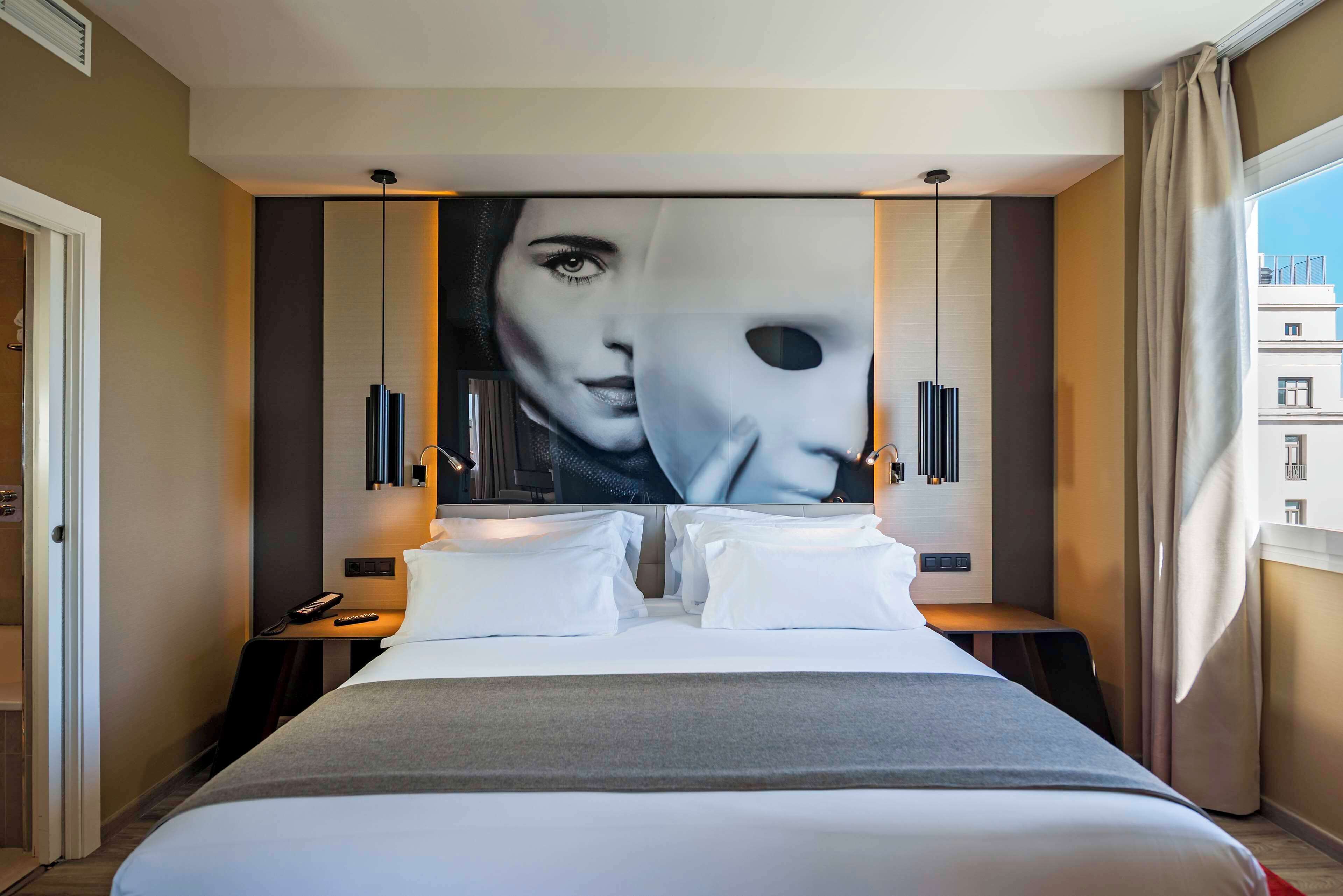 Una habitación de hotel moderna con una cama grande, una mesita de noche, un control remoto y una obra de arte colgada en la pared.
