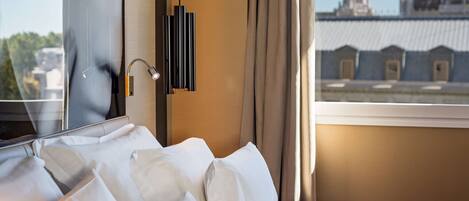 A hotel room with a bed, a window with a view of buildings, and a modern light fixture.