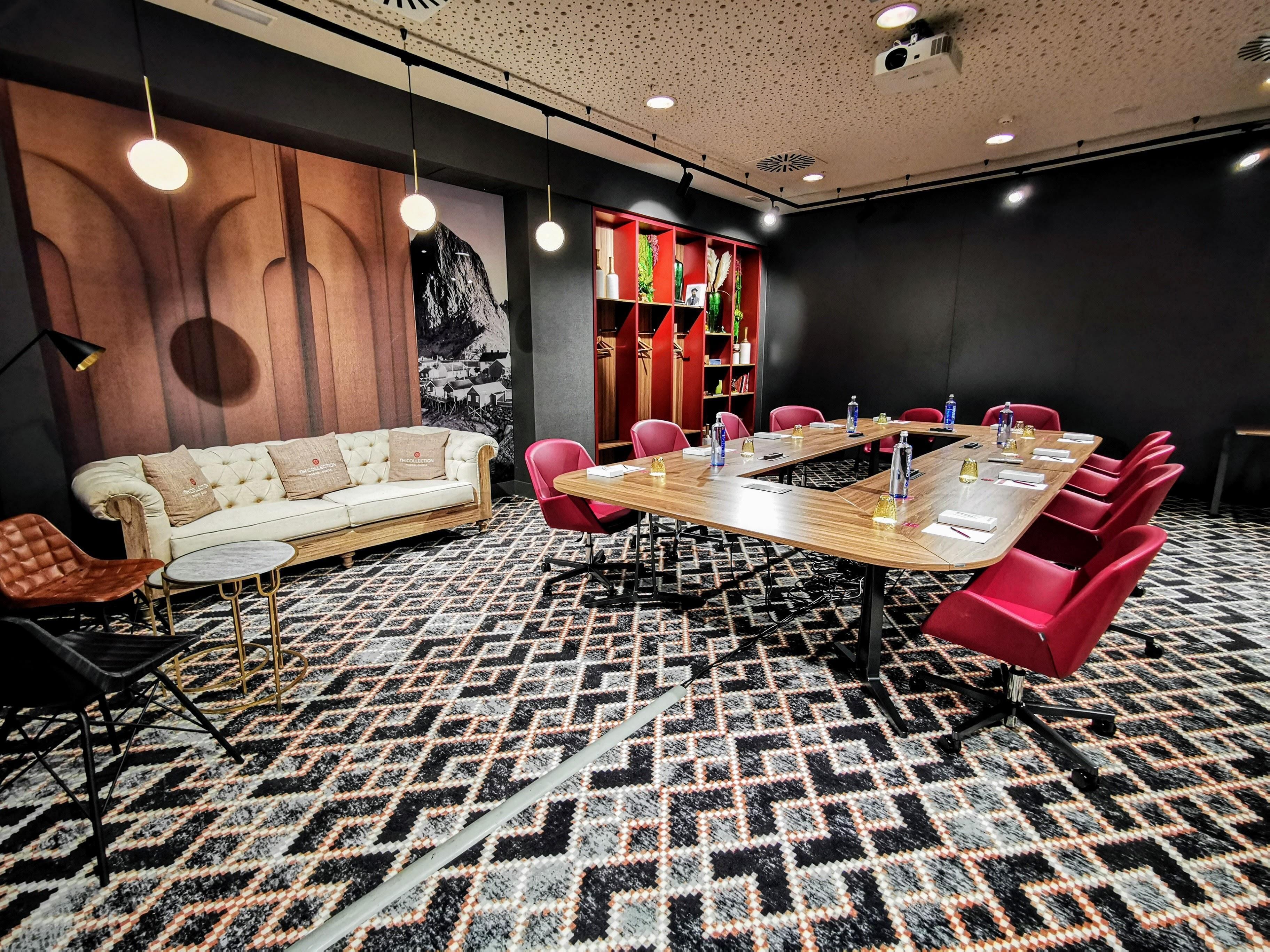 Un salón de conferencias con una gran mesa de madera, sillas rojas, un sofá blanco y una alfombra estampada.