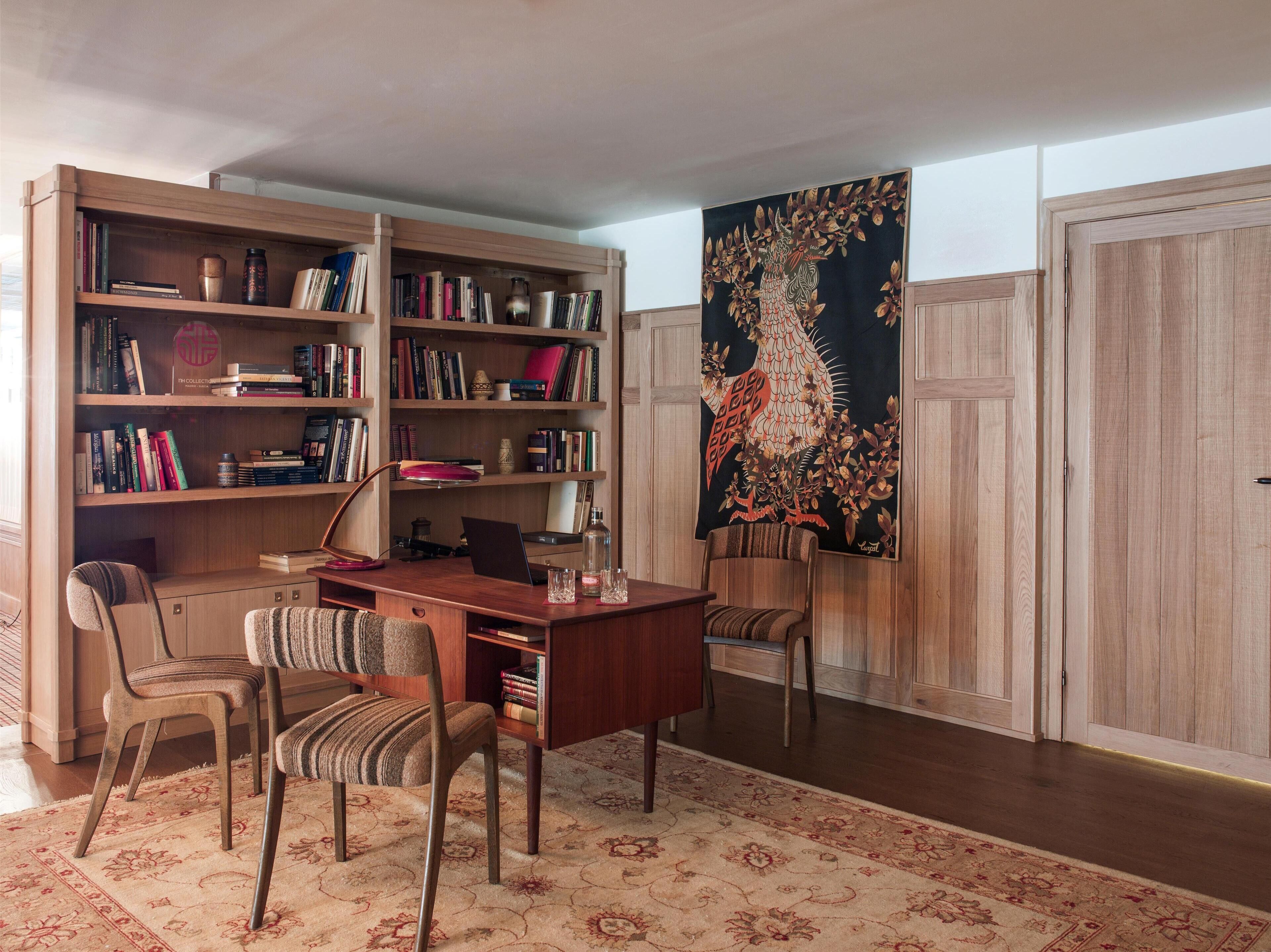 A study room with a wooden desk, two chairs, a bookshelf, and a large tapestry.