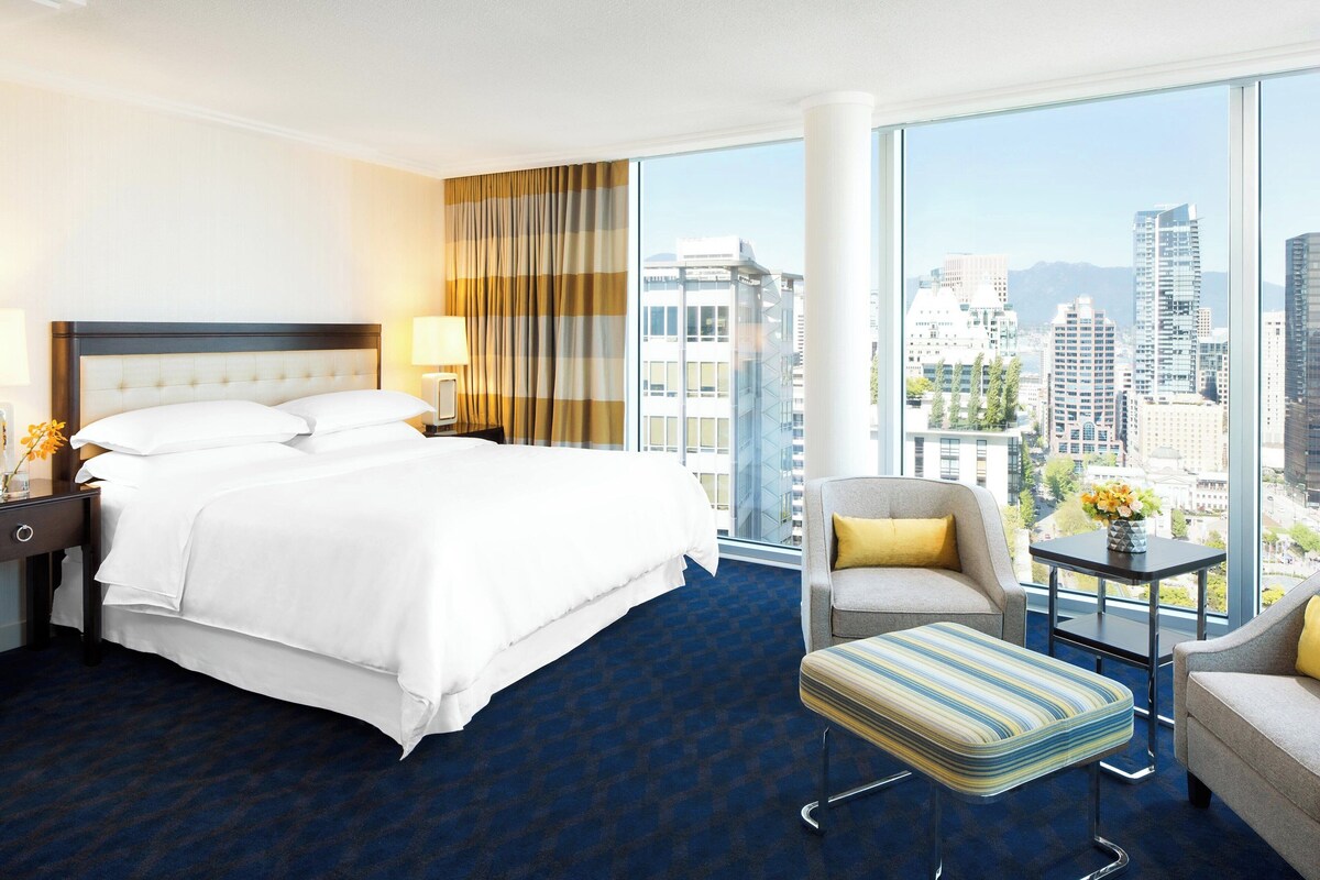 Quarto de hotel com uma cama grande, um espaço de estar com uma poltrona e apoio para os pés, uma mesinha com um vaso, e vista para a cidade através de grandes janelas.