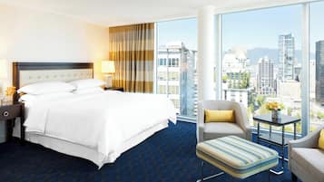 Une chambre d’hôtel dotée d’un grand lit, d’un coin salon avec un fauteuil et un pouf, d’une petite table avec un vase, et offrant une vue sur la ville grâce à de grandes fenêtres.