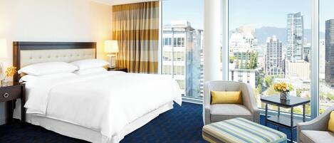 A hotel room with a large bed, a sitting area with a chair and ottoman, a small table with a vase, and a city view through large windows.