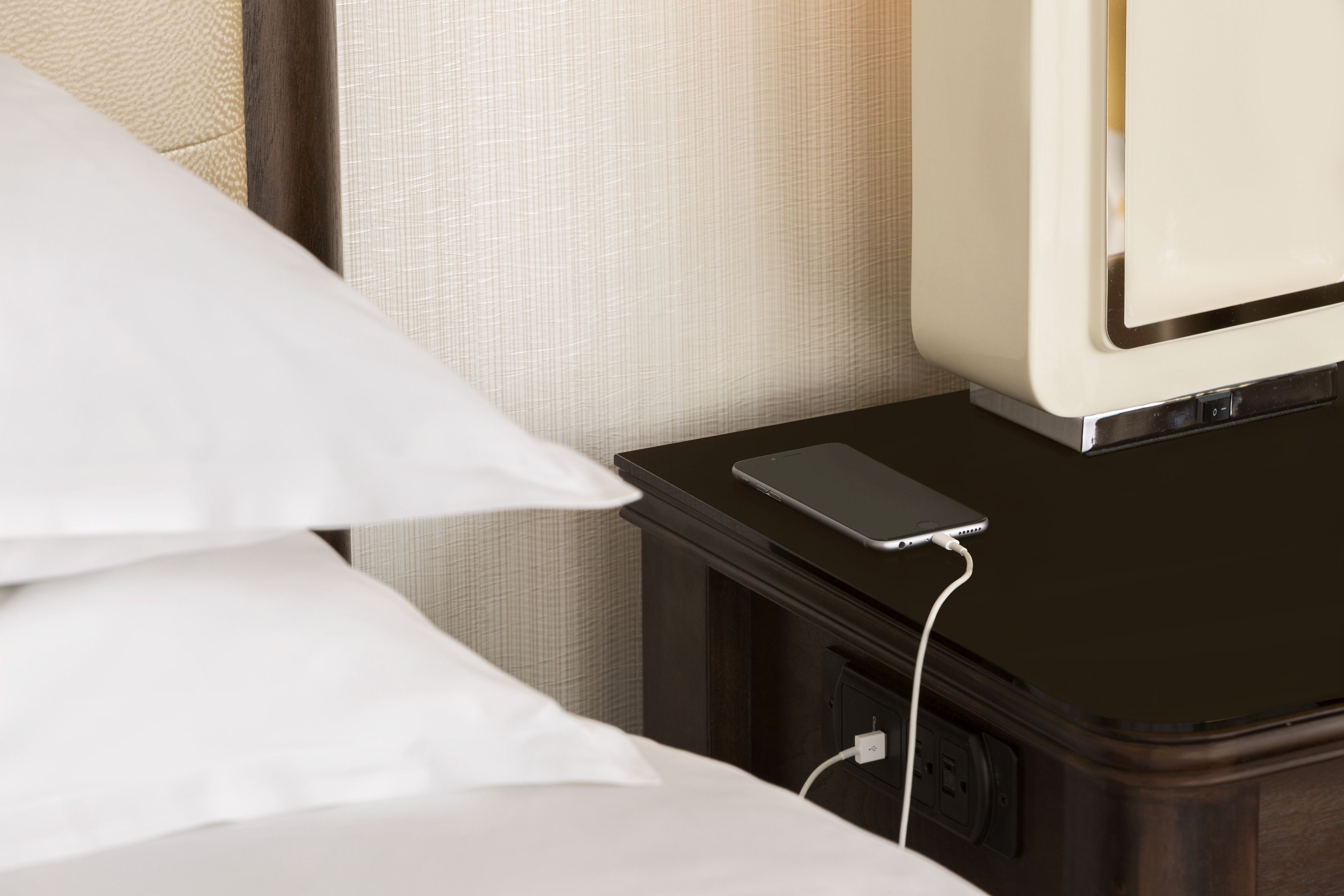 Cama com lençóis brancos, mesa de cabeceira com um smartphone e cabo de carregamento, e parede com textura.