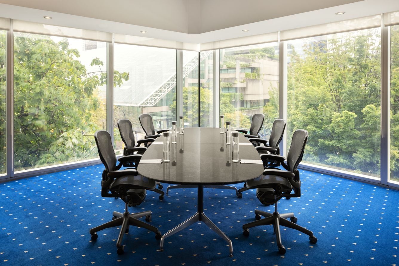 A conference room with a large table surrounded by chairs, large windows with a view of greenery and buildings, and a blue carpet with a pattern.