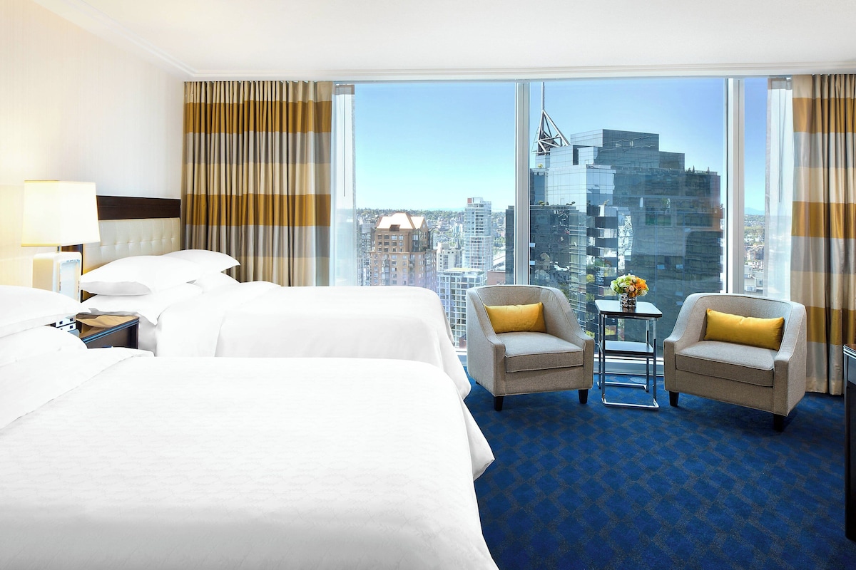 Quarto de hotel com duas camas, um sofá, uma poltrona, uma mesinha com um vaso e uma janela grande com vista para a cidade.
