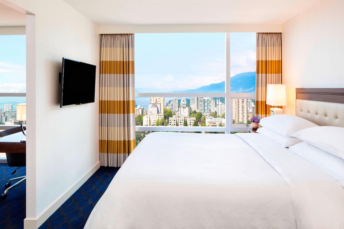 Quarto de hotel com uma cama grande, televisão presa na parede, secretária com cadeira e vista para a cidade através de janelas amplas.
