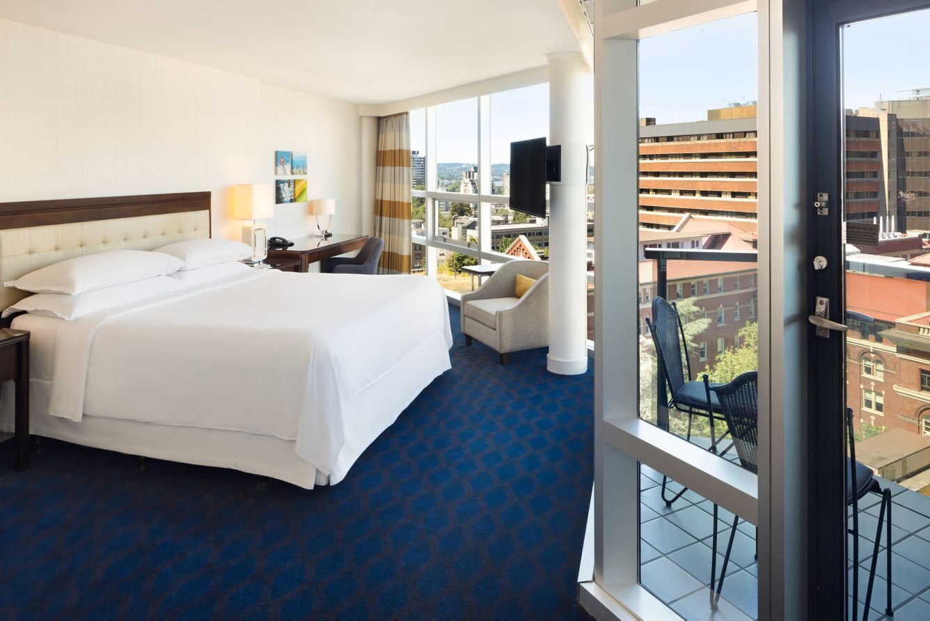 A hotel room with a large bed, a desk with a lamp, a chair, and a view of the city through large windows.