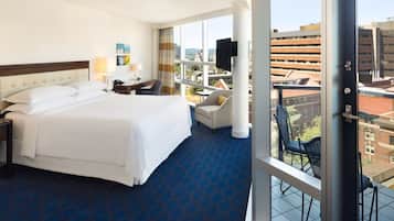 A hotel room with a large bed, a desk with a lamp, a chair, and a view of the city through large windows.