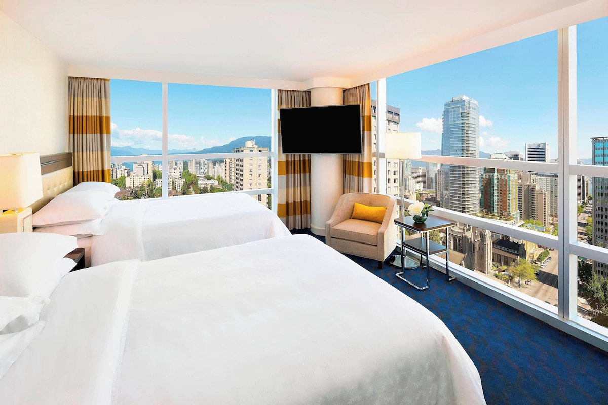 Quarto de hotel com duas camas, uma televisão, uma cadeira e uma mesa pequena.