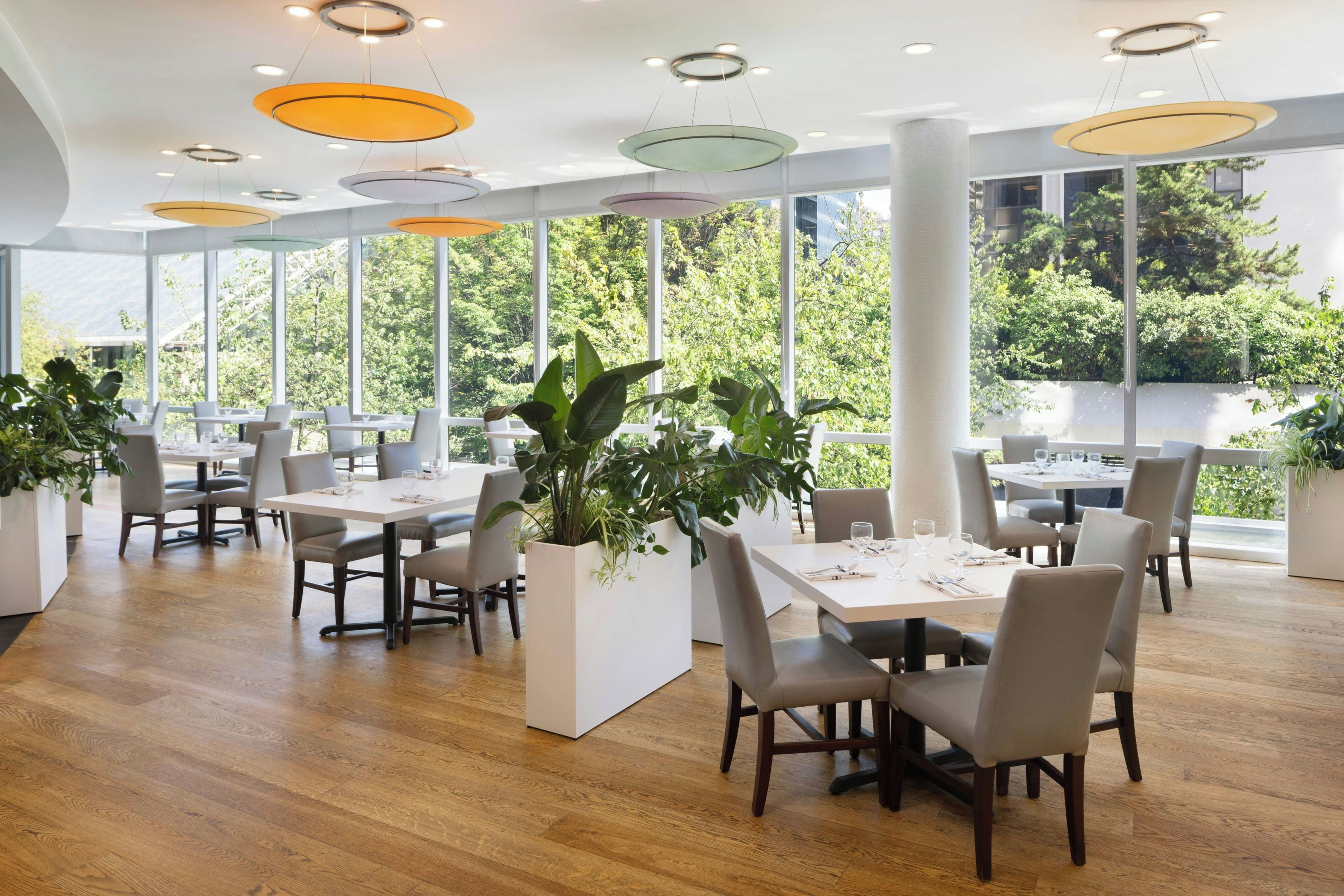 Un área de comedor moderna con piso de madera, mesas y sillas blancas, rodeada de ventanas amplias con vistas a zonas verdes.