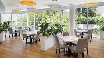 Un área de comedor moderna con piso de madera, mesas y sillas blancas, rodeada de ventanas amplias con vistas a zonas verdes.