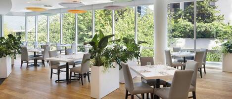 A modern dining area with wooden flooring, white tables, and chairs, surrounded by large windows with a view of greenery.