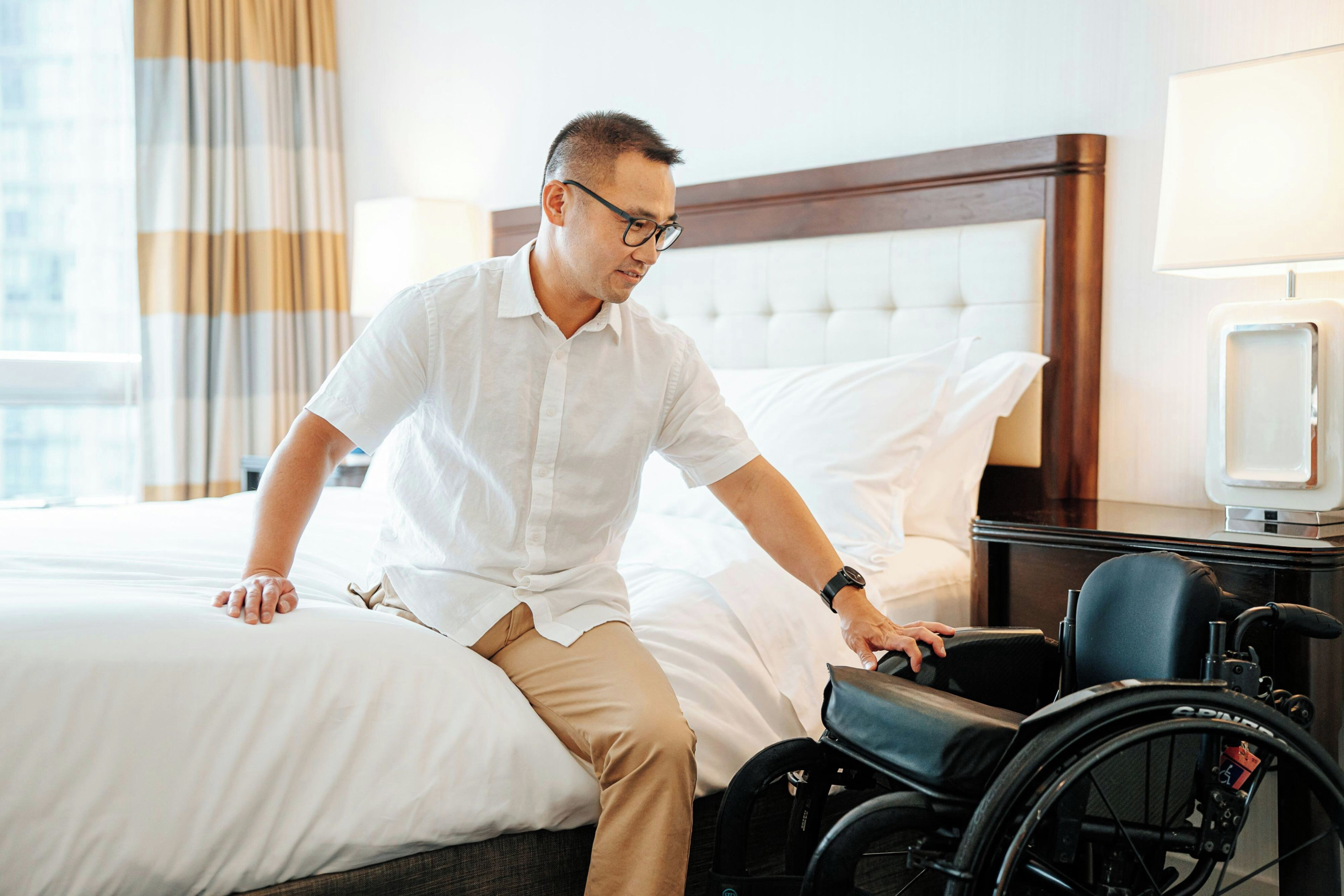 A person sitting on a bed in a hotel room, with a wheelchair beside them.