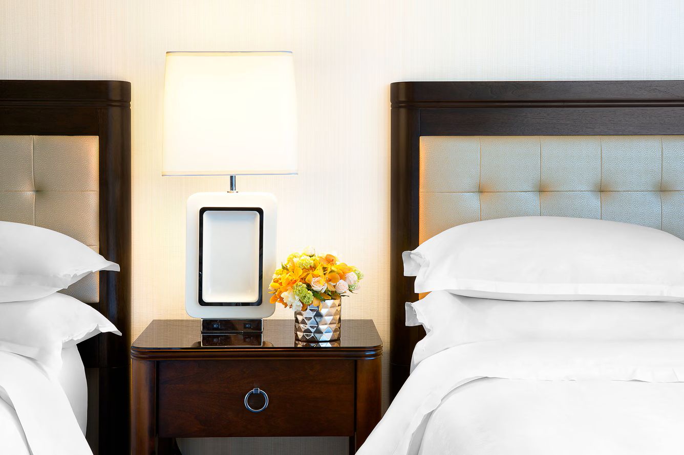 Quarto de hotel com duas camas, uma mesa de cabeceira com abajur e um vaso com flores.