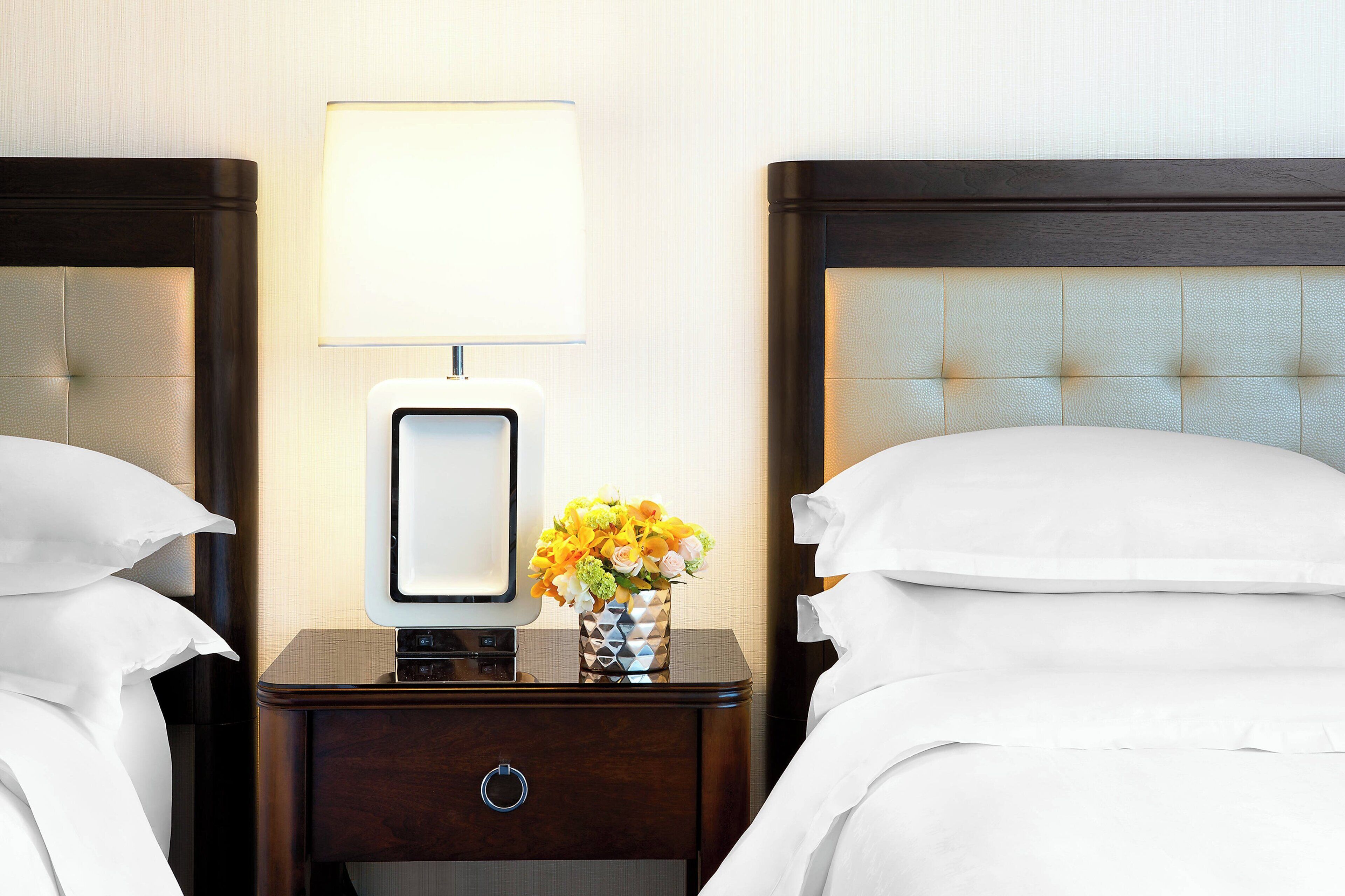 Habitación de hotel con dos camas, una mesita de noche con lámpara y un jarrón con flores.