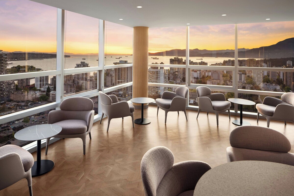 Un salón moderno con varias zonas de estar, amplios ventanales con vistas a la ciudad y un suelo de madera con patrón de espiga.