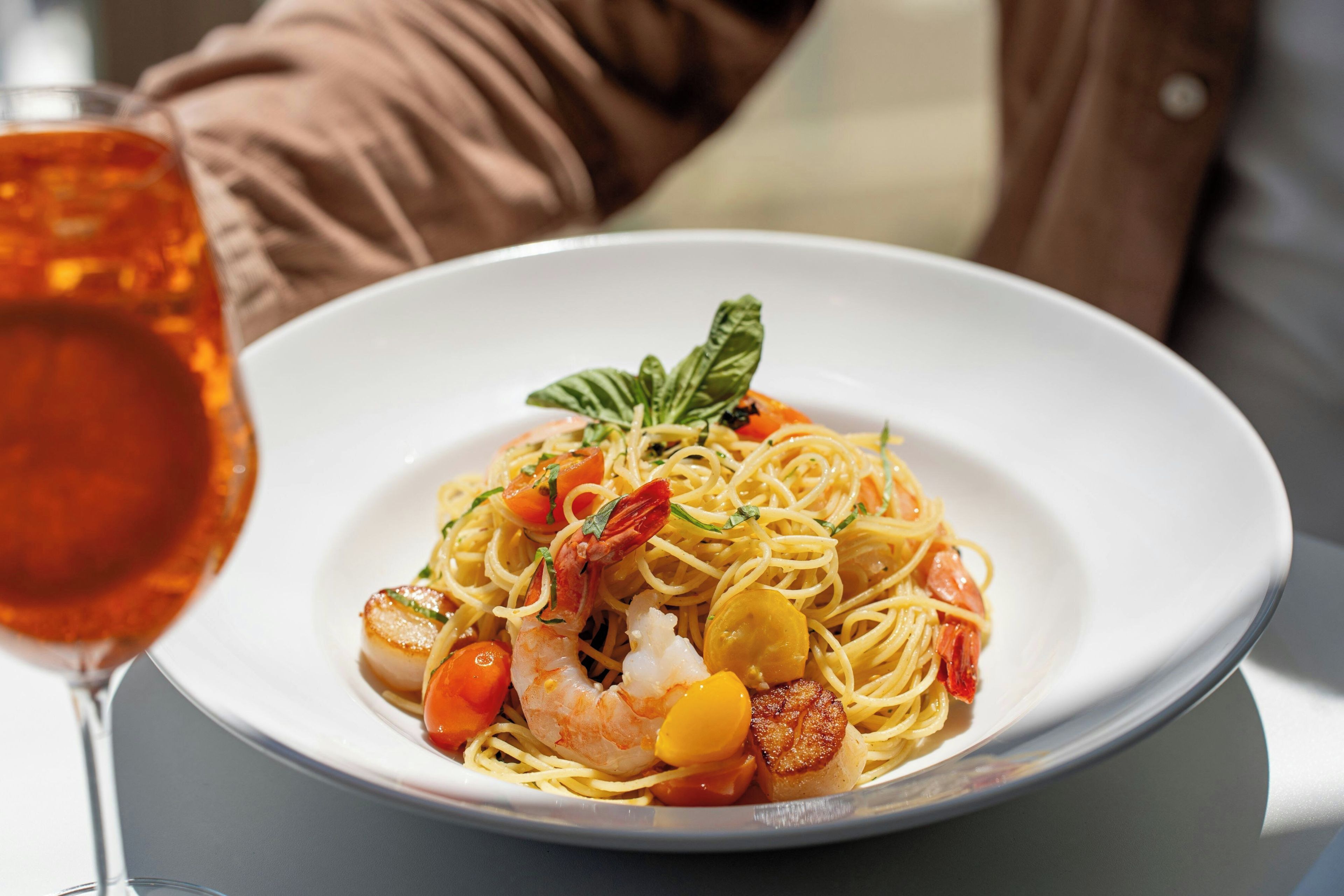 A plate of pasta with shrimp and vegetables, a glass of red wine, and a brown cushion in the background.