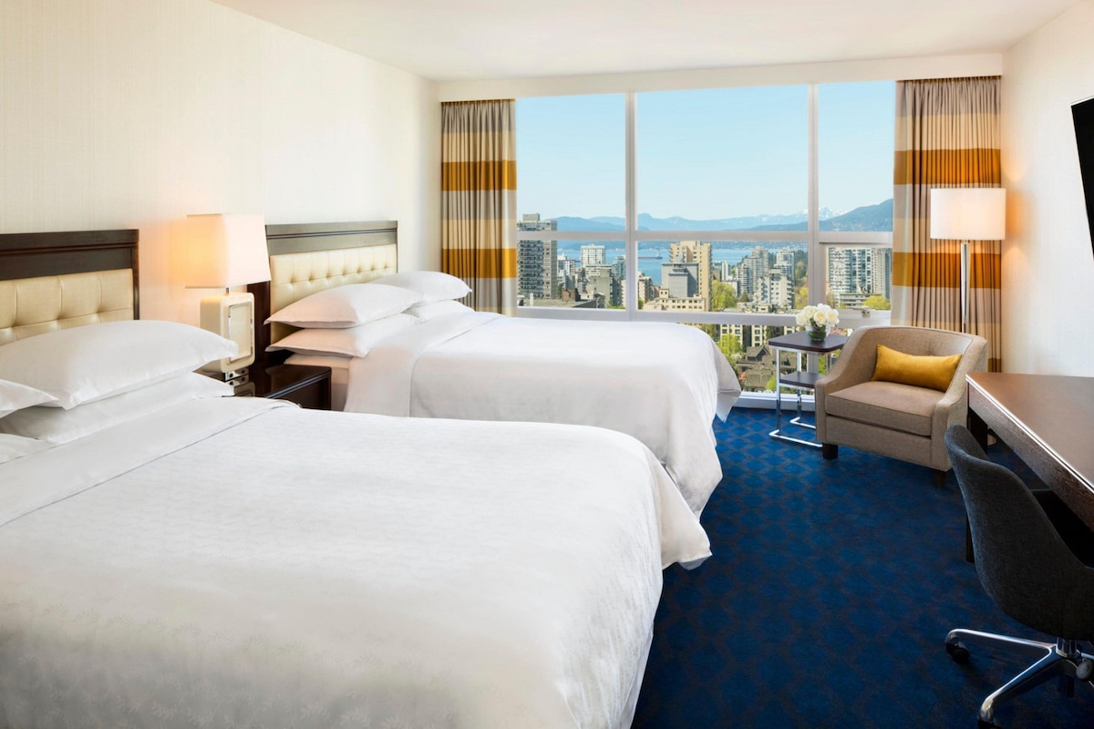 Quarto de hotel com duas camas, uma escrivaninha, uma cadeira e vista para a paisagem urbana.