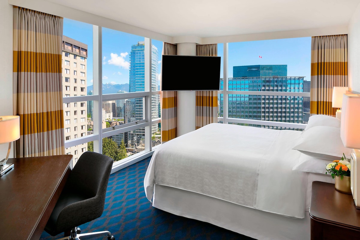 Quarto de hotel com uma cama grande, uma mesa, uma cadeira, uma televisão e vista para a cidade.