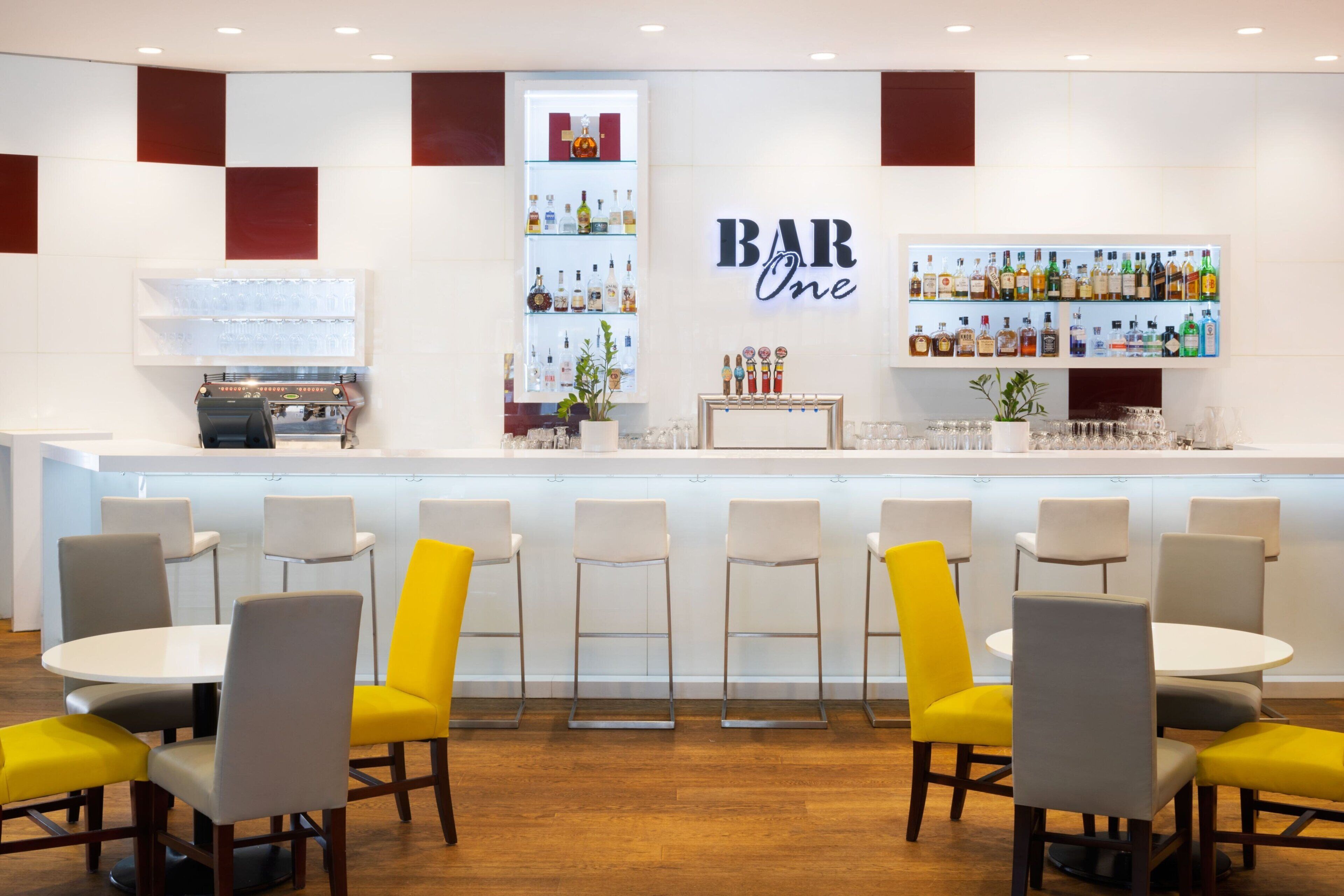 Uma área de bar com balcão branco, prateleiras com garrafas de bebidas e banquetas.
