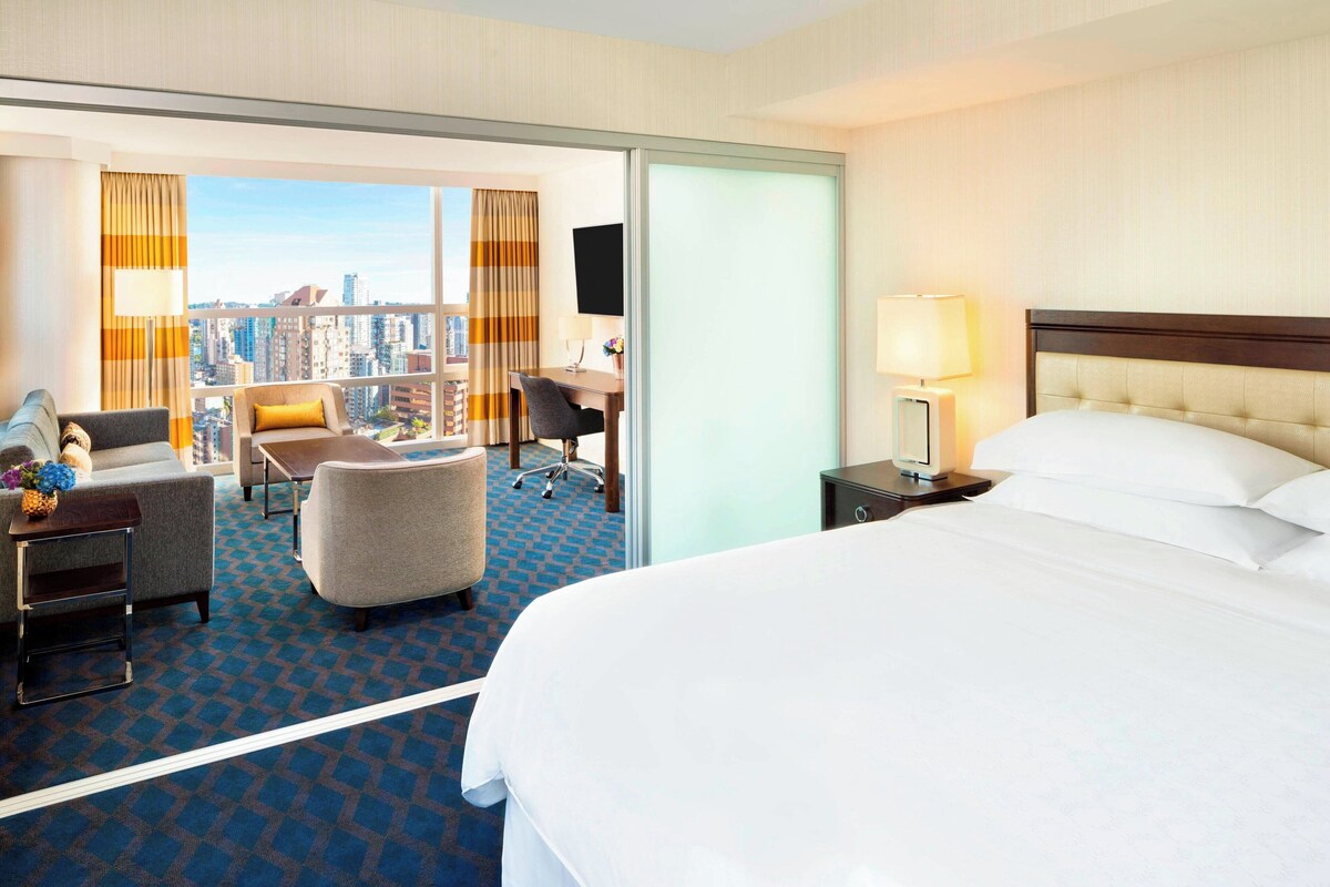 Quarto de hotel com uma cama grande, uma escrivaninha, uma cadeira e um sofá. Vê-se a cidade pela janela.
