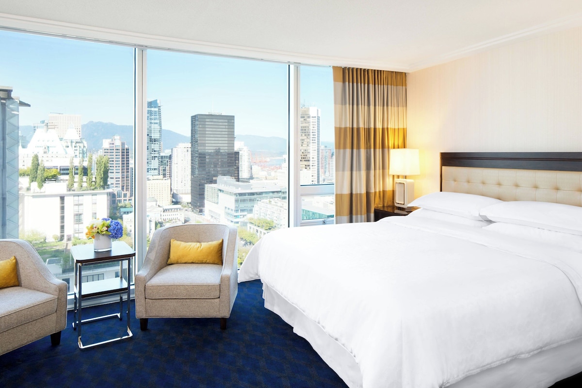 Quarto de hotel com uma cama grande, duas poltronas, uma mesa pequena com um vaso e vista para a paisagem urbana através de amplas janelas.