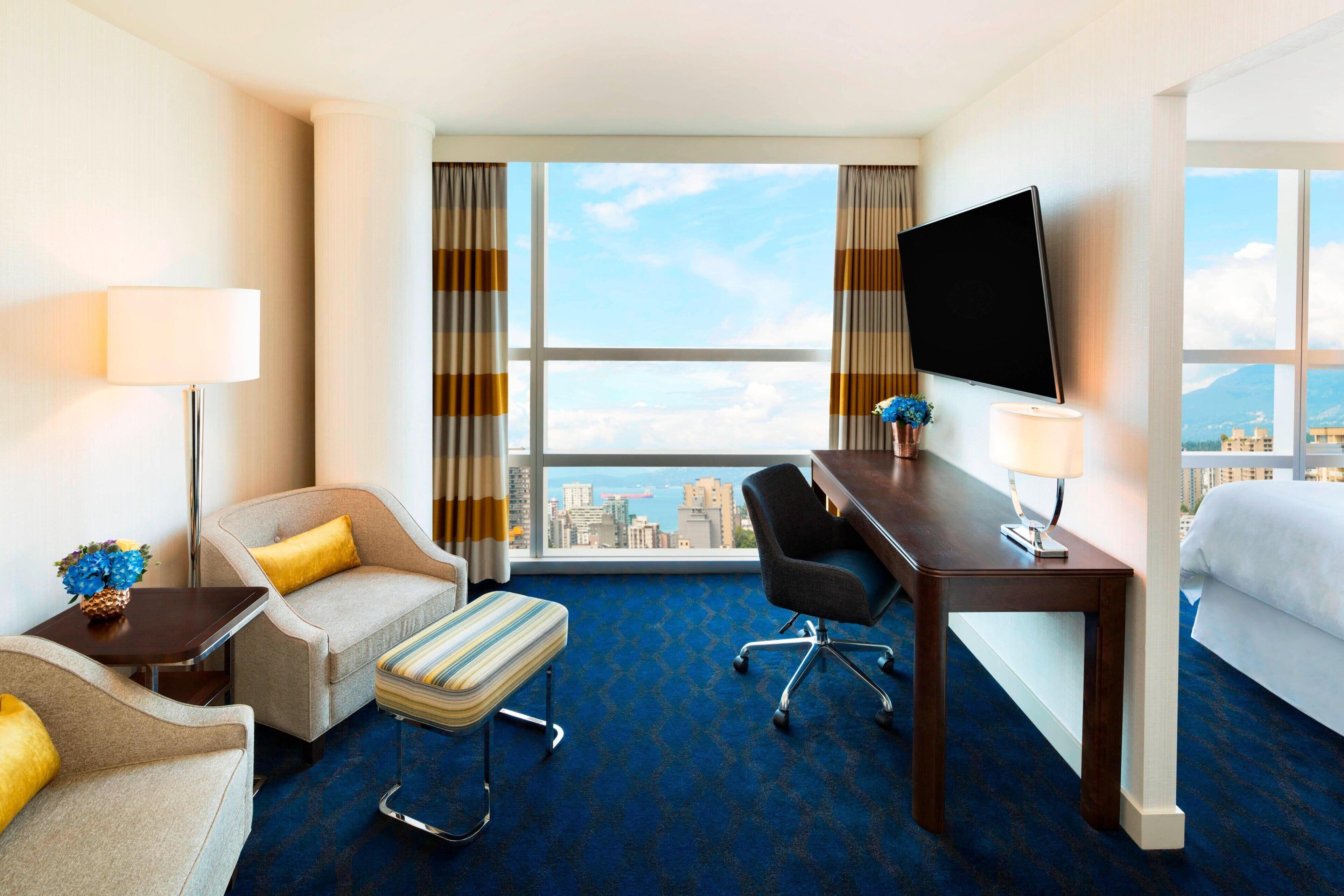 A hotel room with a large window, a flat-screen TV, a desk, a chair, a sofa, and a bed.