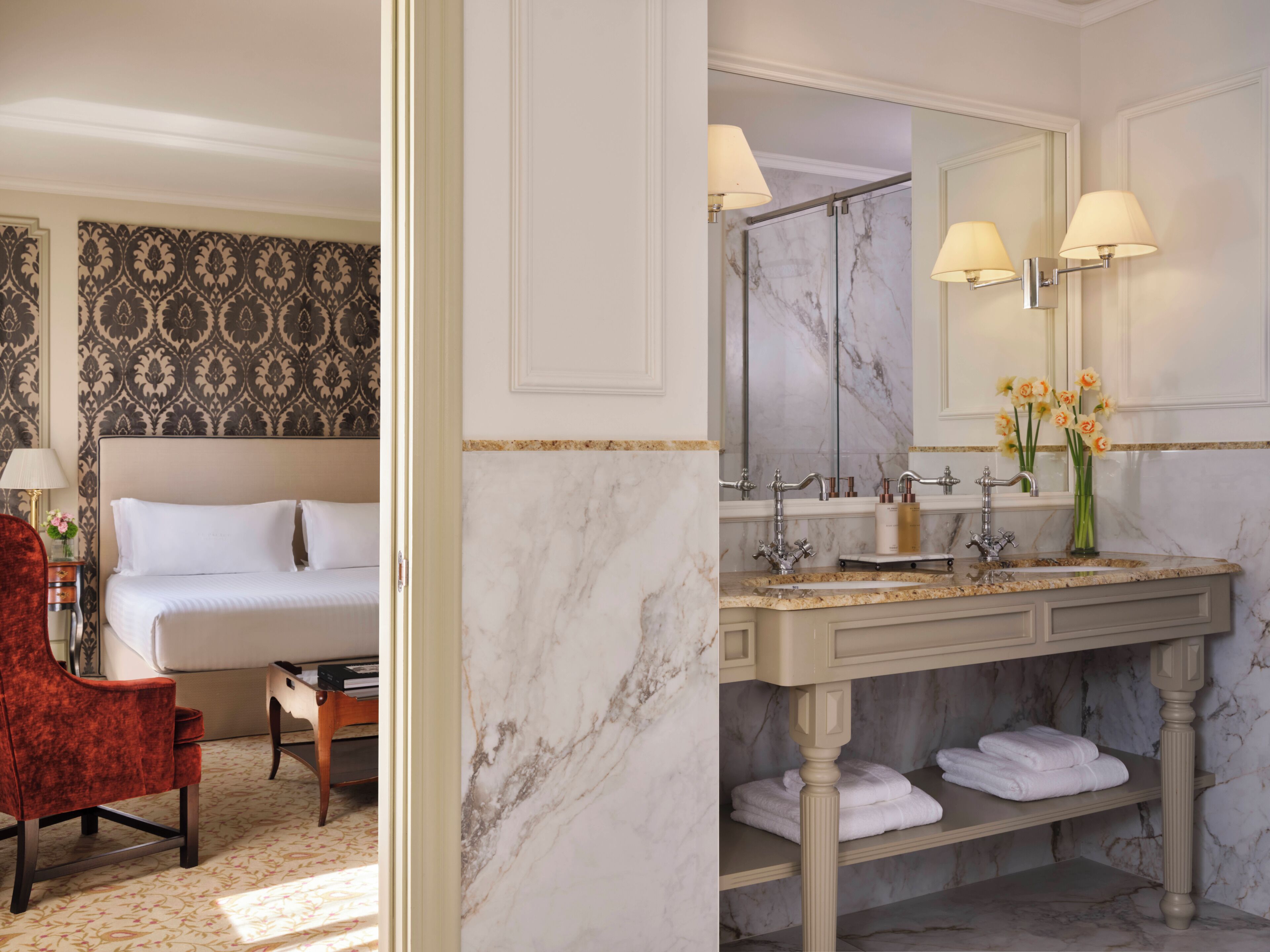 A hotel room with a marble bathroom, a bed with white linens, a red chair, and a vanity with a mirror.